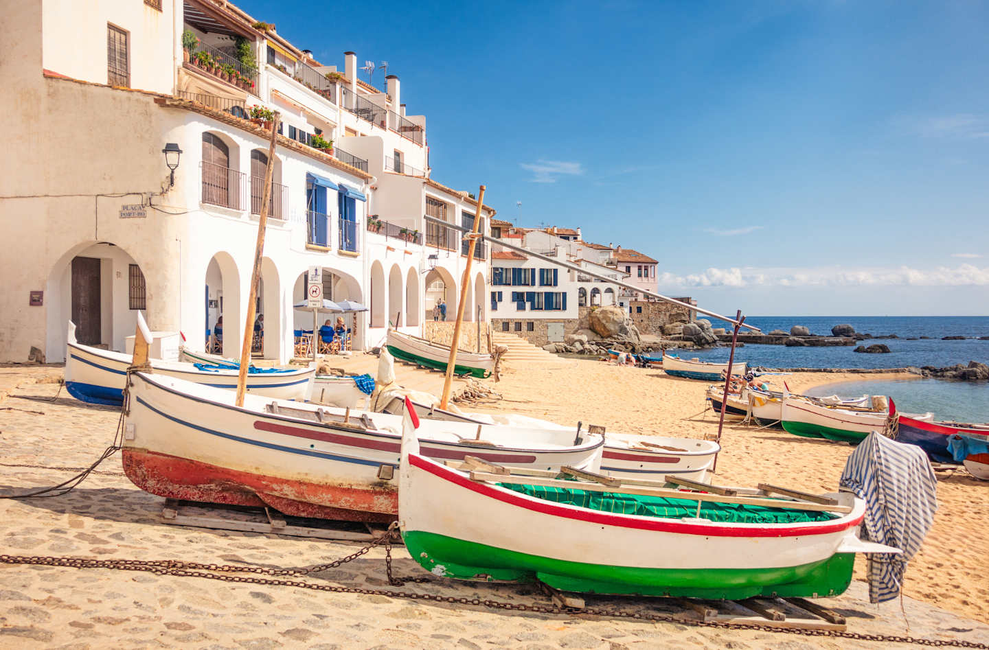 A colorful array of traditional fishing boats rests on the sandy beach, surrounded by charming Mediterranean-style buildings and a clear blue sky.