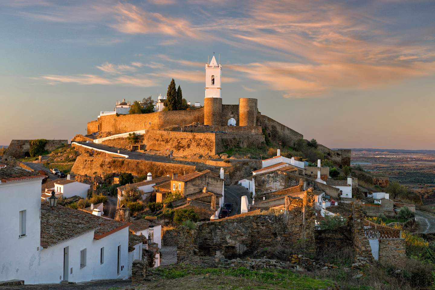 A picturesque hilltop town with a medieval castle and church, surrounded by colorful houses and set against a dramatic sky with vibrant clouds at sunset.
