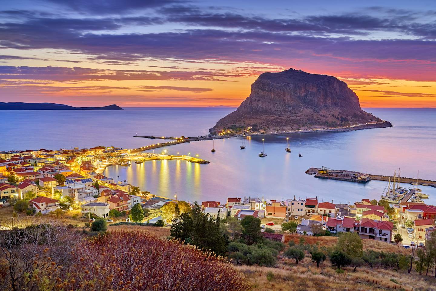A picturesque coastal town nestled at the base of a towering rock formation, with a vibrant sunset illuminating the scene and boats dotting the calm waters.