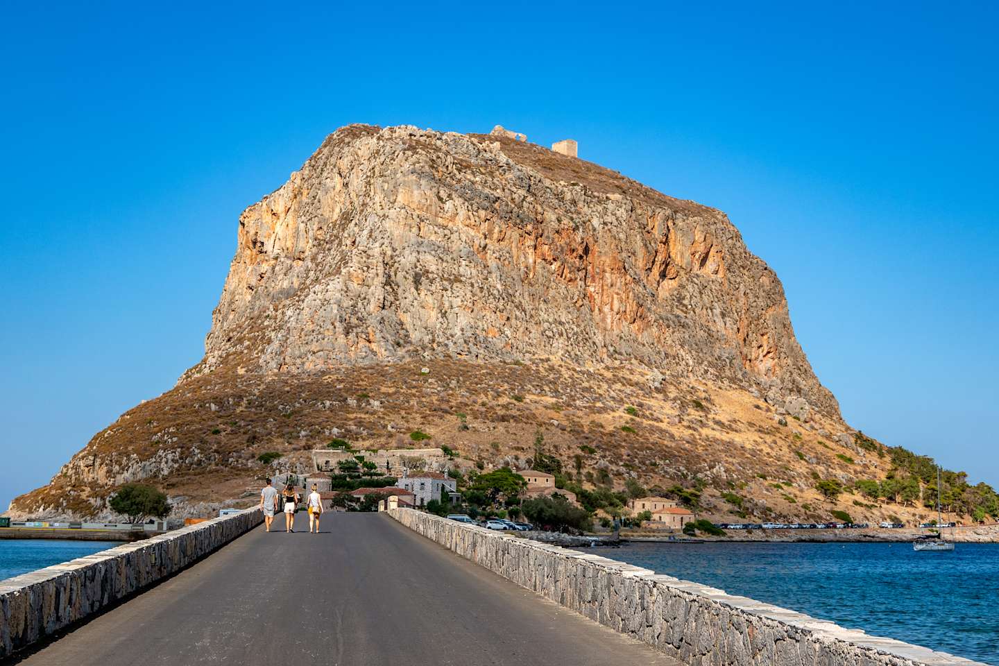 A long, narrow bridge leads to a towering, rocky mountain with a castle or fortress perched atop it, set against a vibrant blue sky.