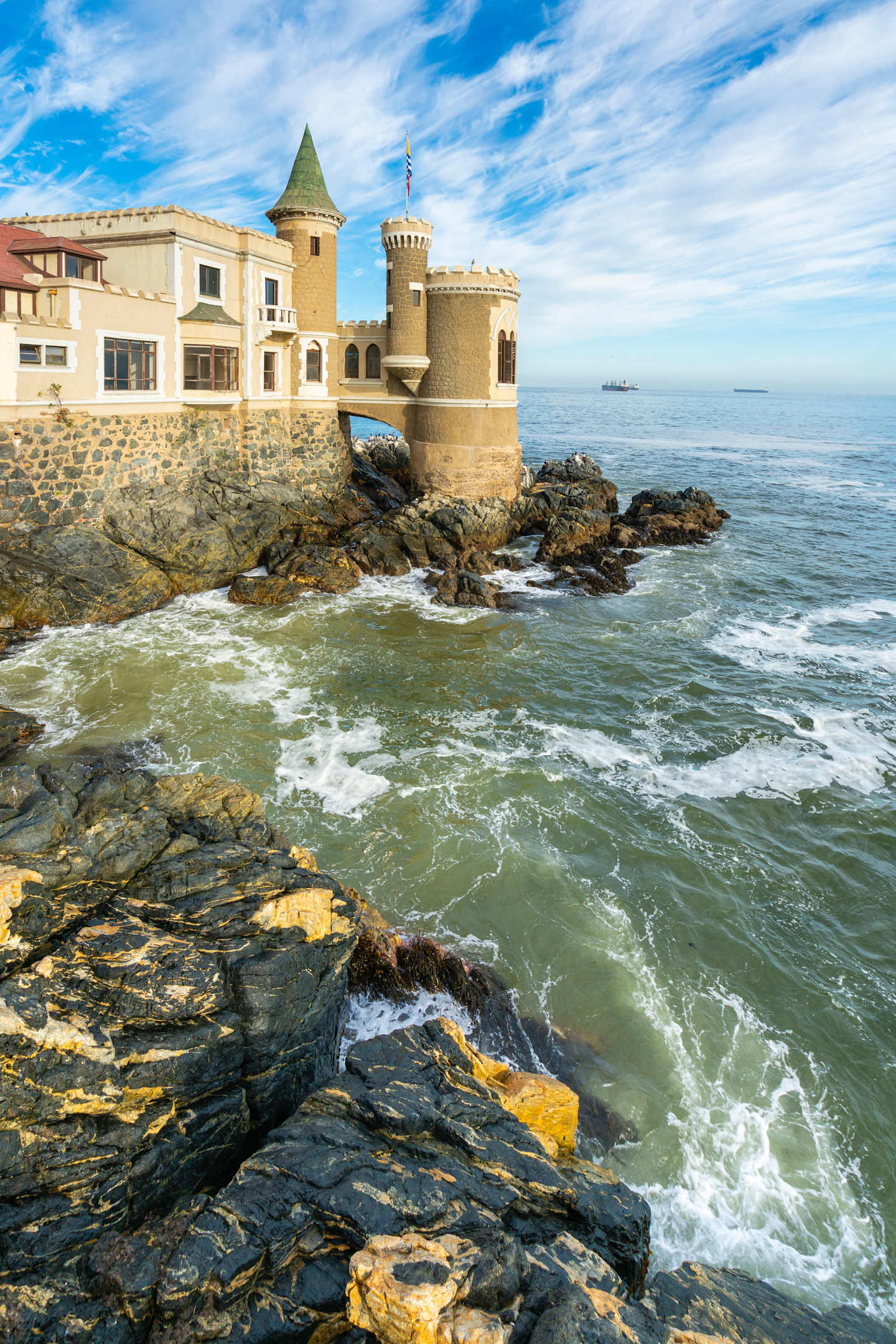 A picturesque castle perched atop rocky cliffs, surrounded by crashing waves and a vibrant blue sky with fluffy clouds.
