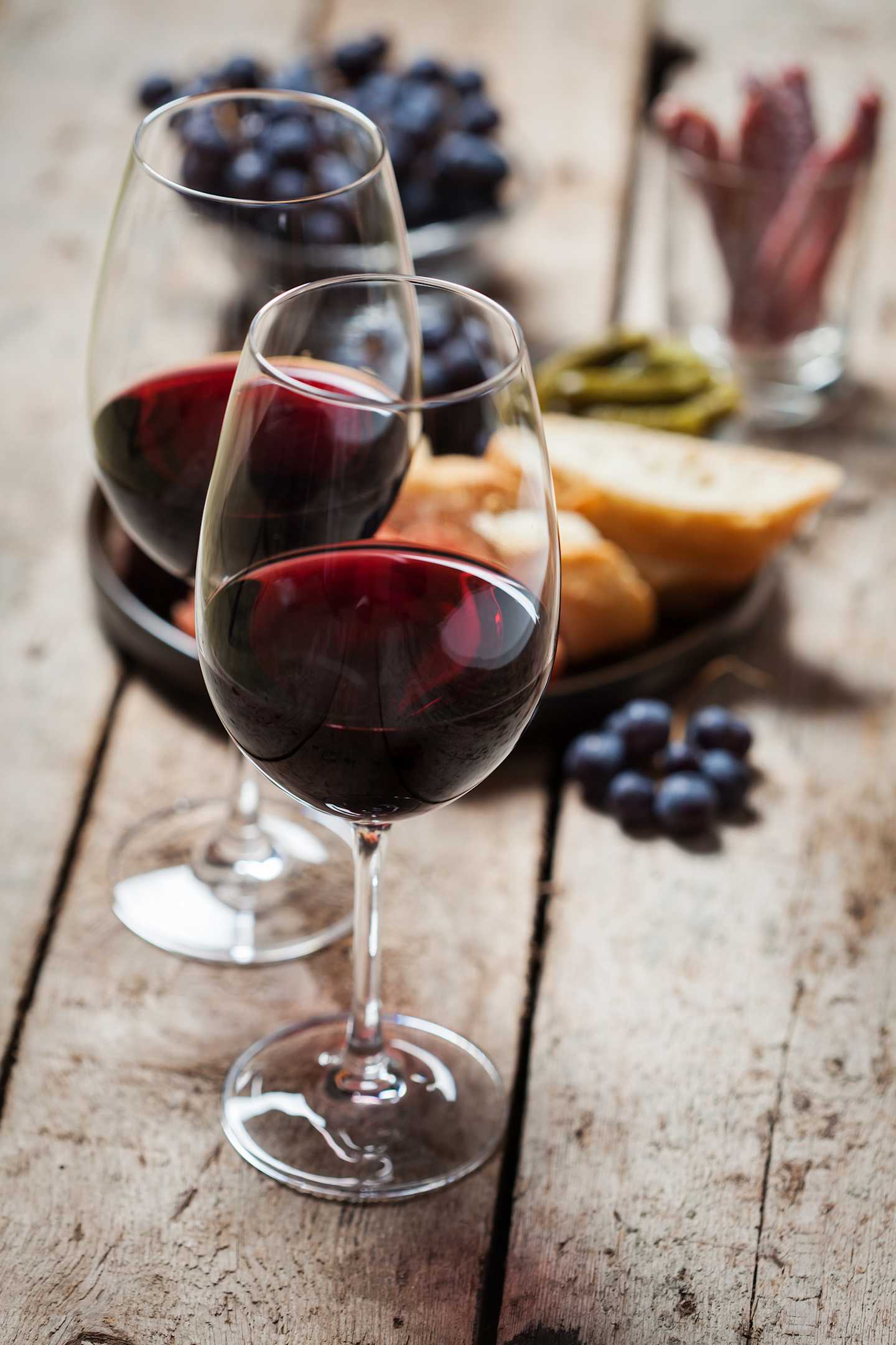 Two glasses of red wine on a wooden table, surrounded by various fruits and snacks, including grapes, cheese, and dried meat.