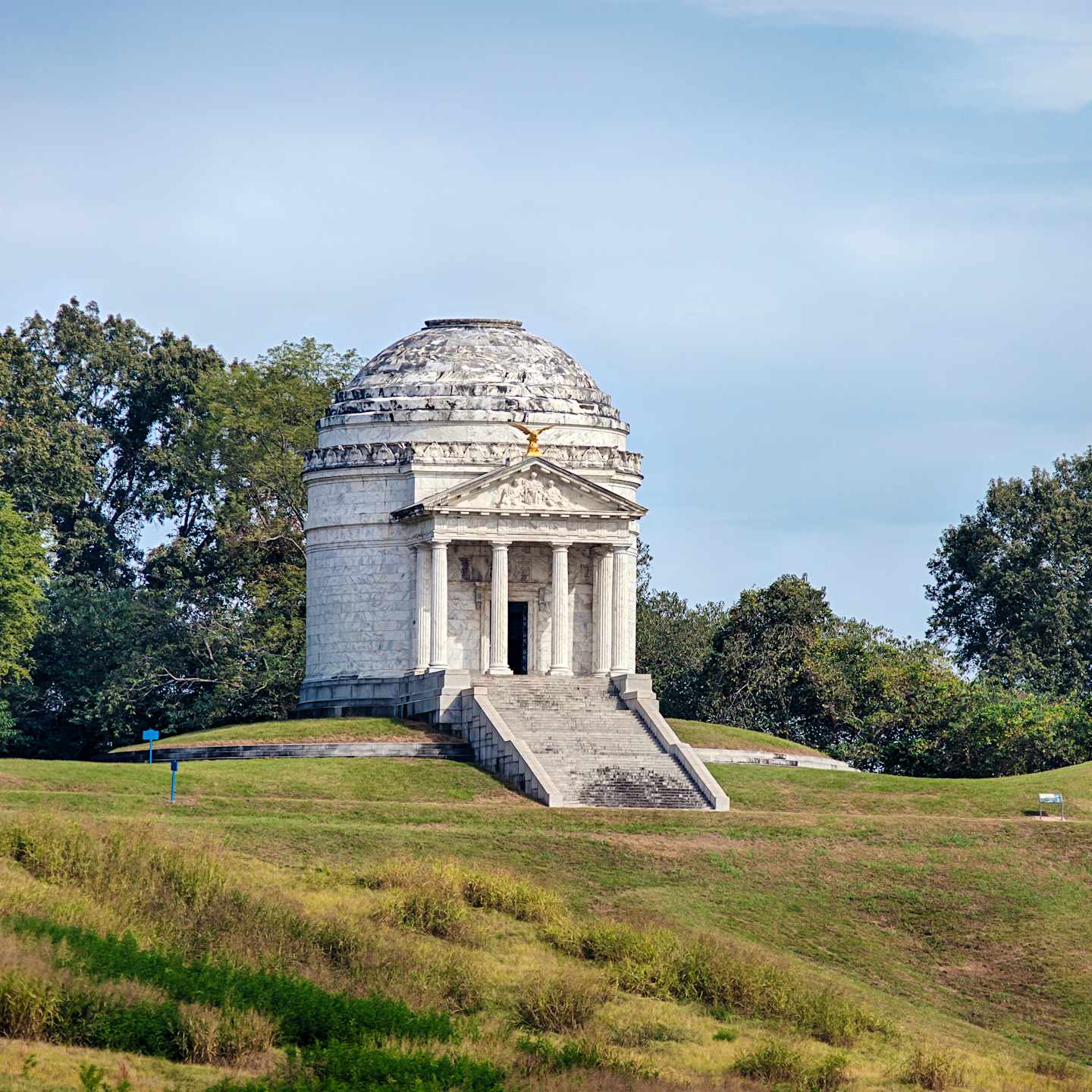 A large, domed monument stands atop a grassy hill, surrounded by trees and a clear blue sky.