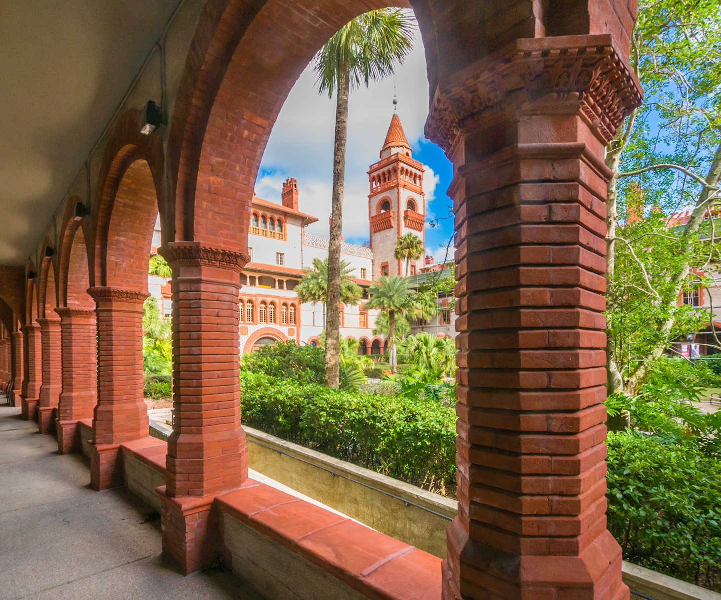 A brick archway frames a lush, verdant courtyard with a towering clock tower in the background, creating a picturesque and historic architectural scene.