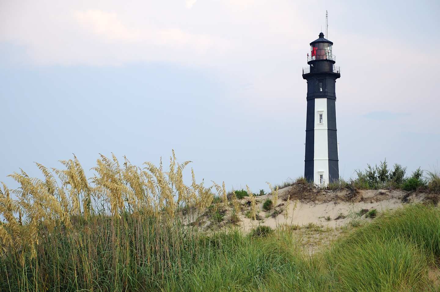 A tall, black-and-white lighthouse stands prominently against the sky, surrounded by tall grass and sand dunes in the foreground.