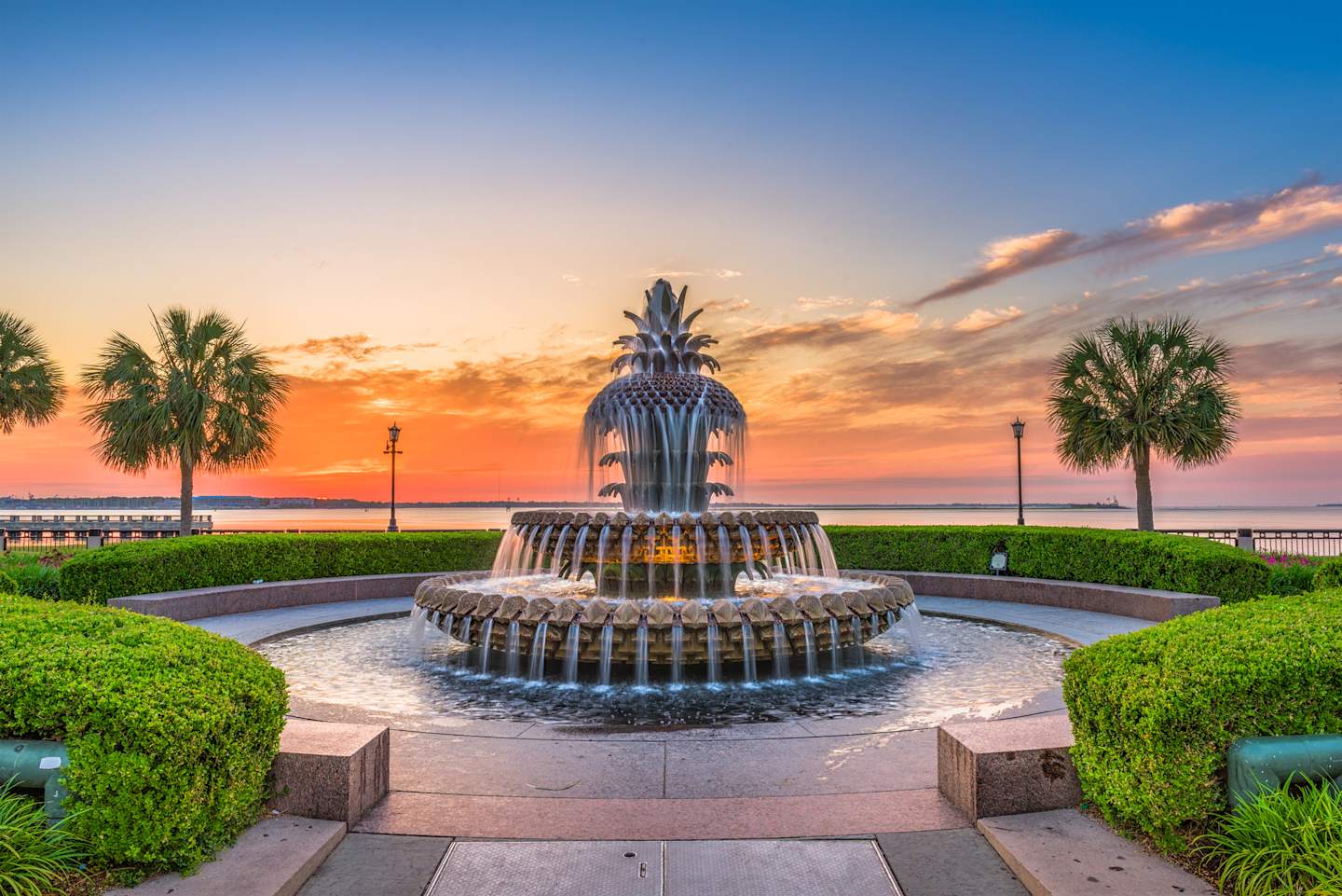 A beautiful pineapple-shaped fountain stands in the foreground, surrounded by lush greenery, against a vibrant sunset sky with palm trees in the background.