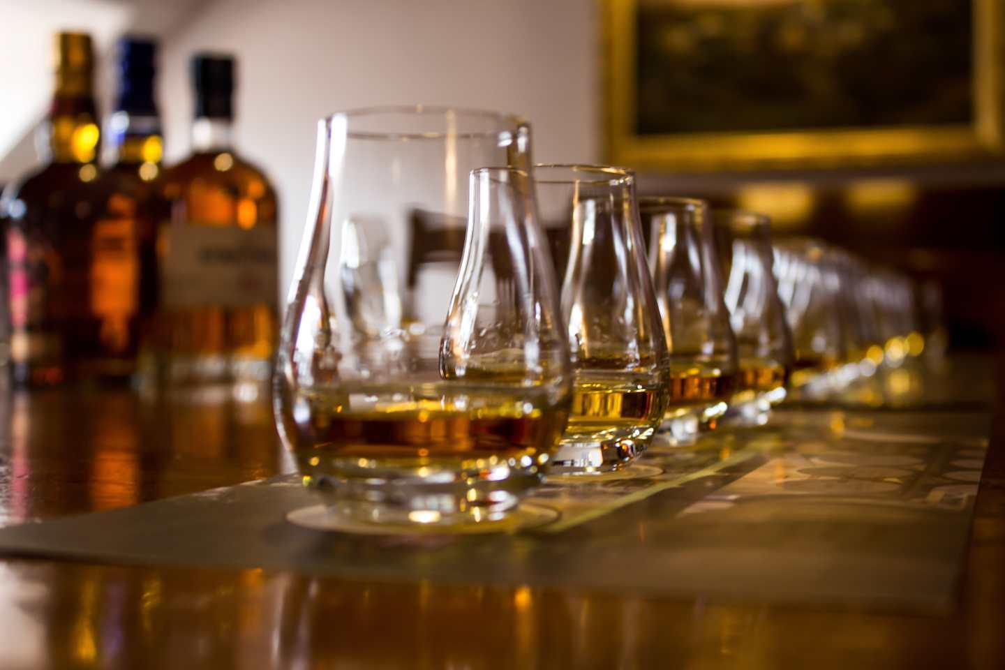 A row of whiskey glasses filled with amber liquid, with bottles of whiskey visible in the background on a wooden surface.