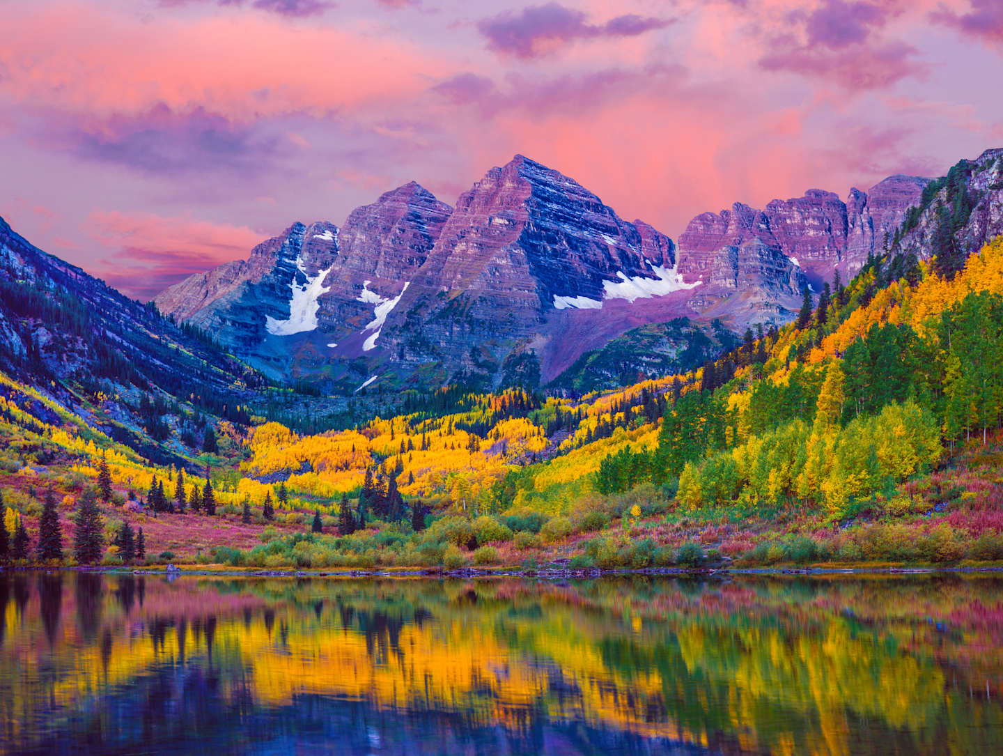 Majestic snow-capped mountains rise against a vibrant sky, their reflection mirrored in the serene lake below, surrounded by a vibrant display of autumn foliage.