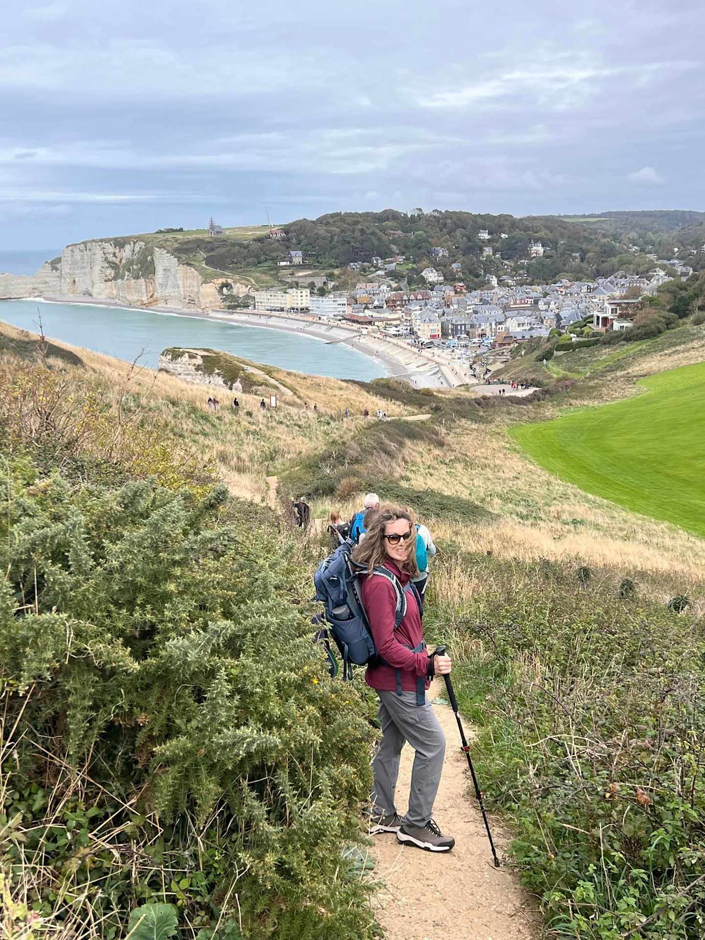 A hiker with a backpack walks along a trail overlooking a coastal town nestled between hills and the ocean.