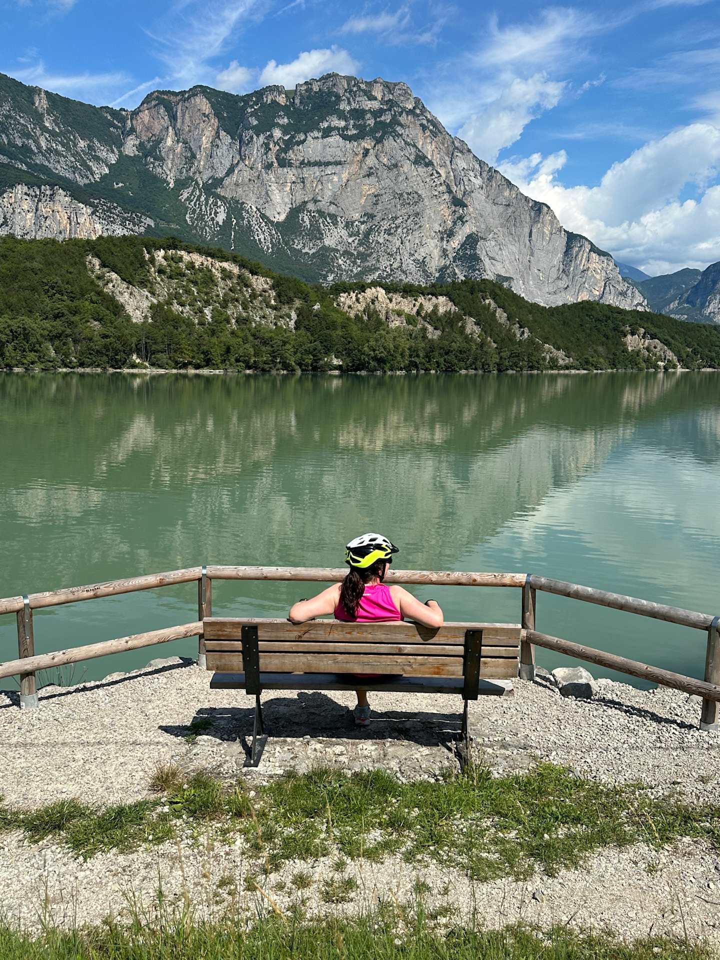 A person in a pink shirt sits on a bench overlooking a serene lake surrounded by majestic mountains in the background.
