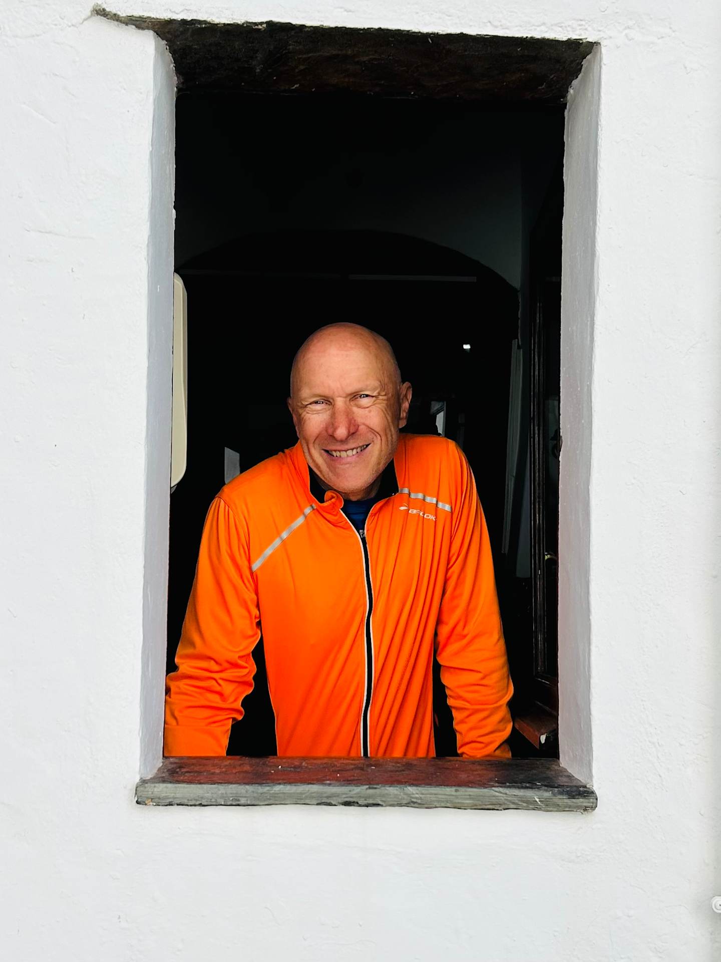A smiling man in an orange jacket stands in a doorway against a dark background.