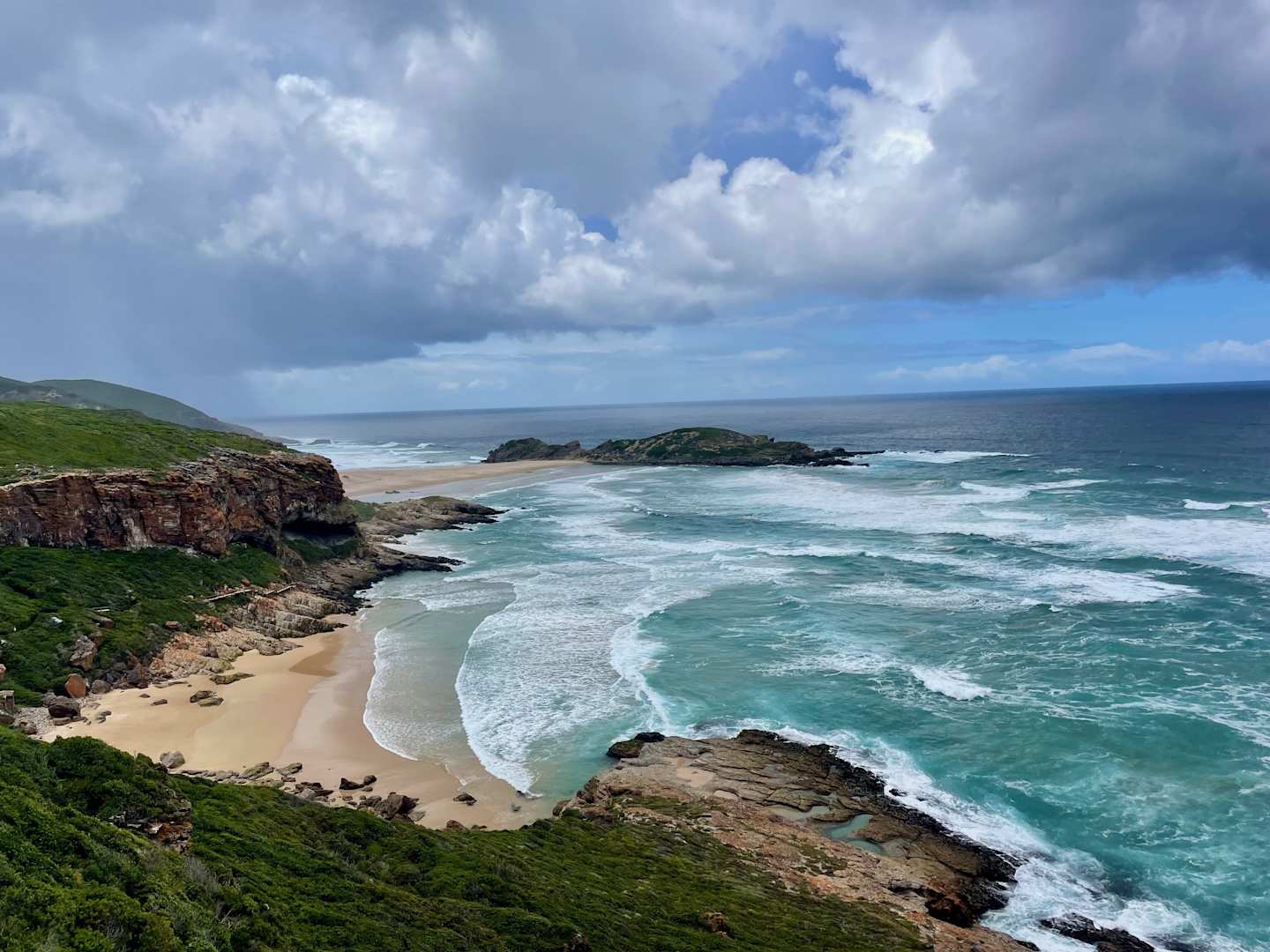 A rugged, rocky coastline with a sandy beach and crashing waves, set against a backdrop of stormy clouds and a vast, blue ocean.