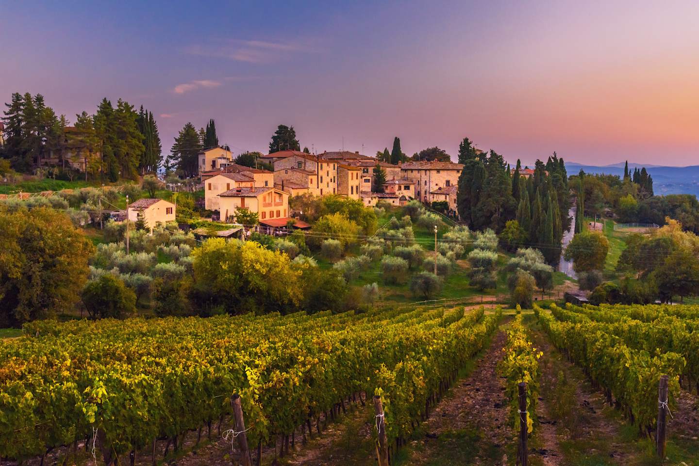 A picturesque Italian village nestled among rolling vineyards, with a stunning sunset backdrop illuminating the charming architecture and lush greenery.