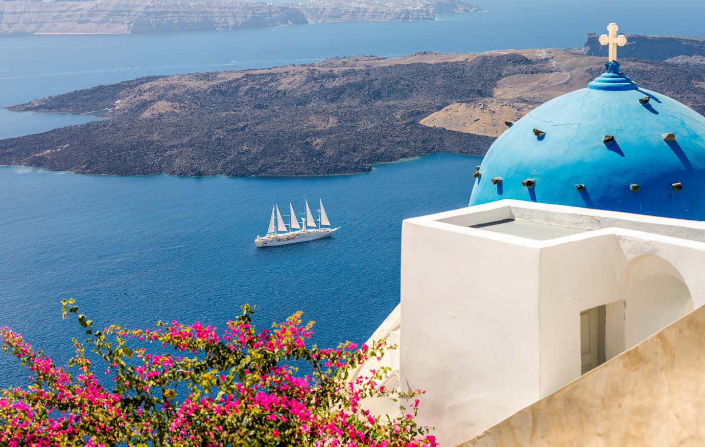 A picturesque view of a blue-domed church overlooking a serene, azure sea with a sailboat in the distance, surrounded by vibrant pink flowers in the foreground.