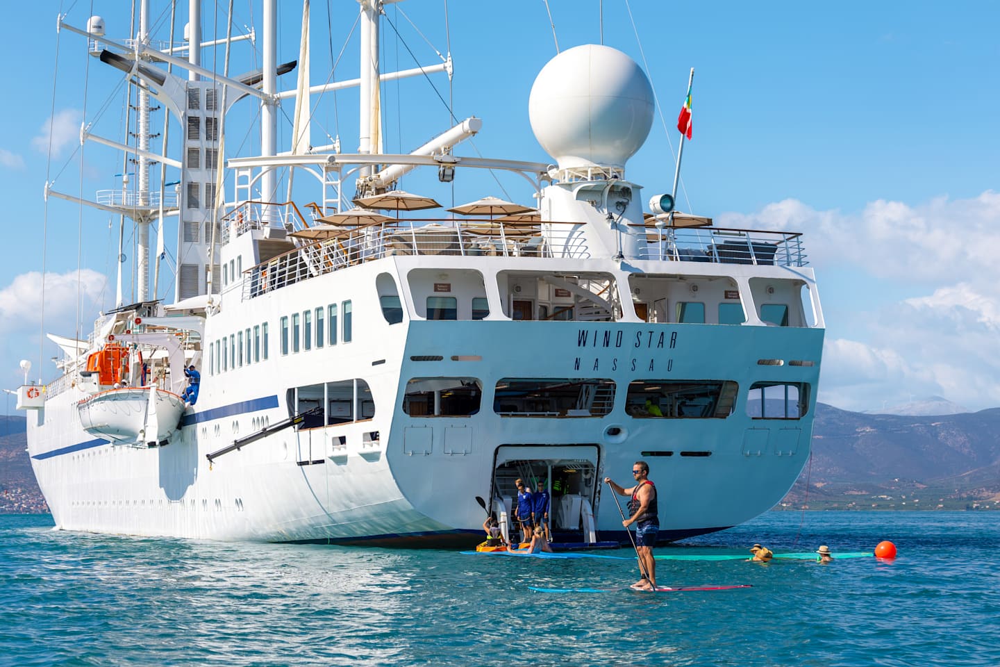 A large white cruise ship with various antennas and equipment on its deck is anchored in a body of water, surrounded by people swimming and enjoying the sunny day.