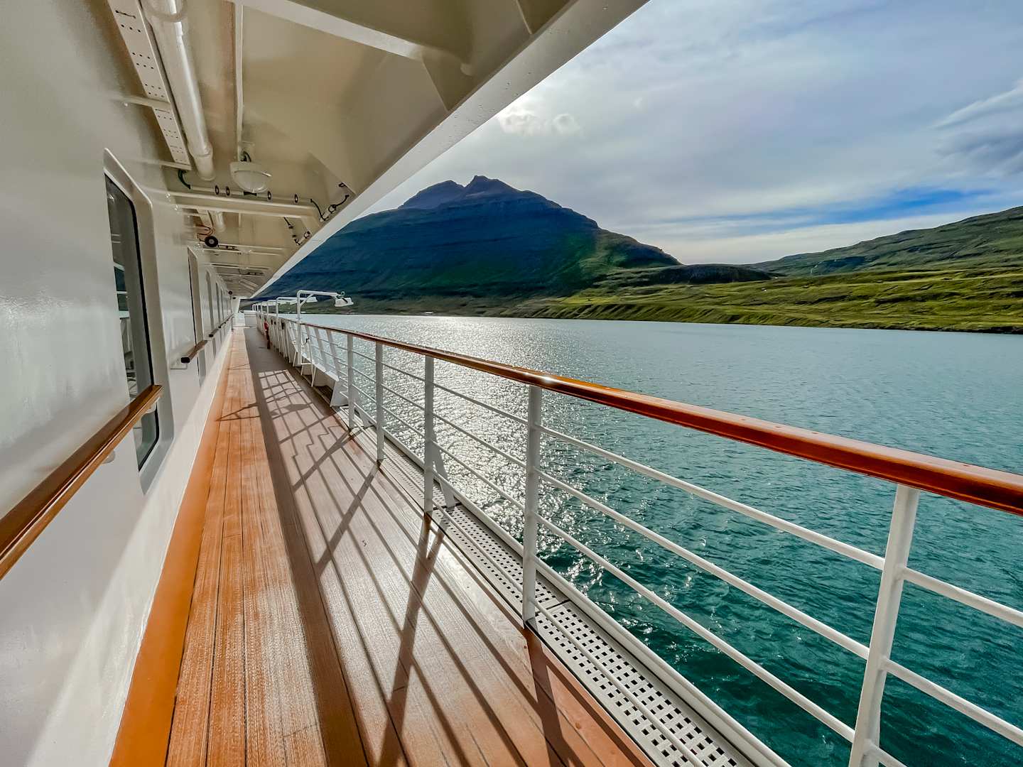 A scenic view from the deck of a ship, with a mountainous landscape in the background and a wooden deck with metal railings in the foreground, overlooking the calm, blue waters.