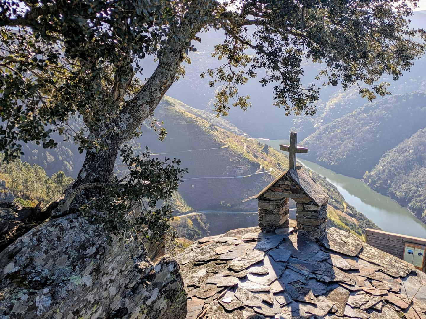 A stone chapel with a cross atop sits on a rocky outcrop, overlooking a serene lake surrounded by lush, forested mountains in the distance.