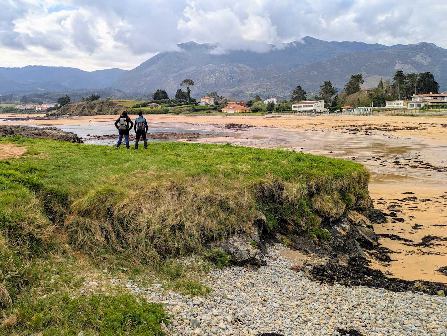 A scenic coastal landscape with a grassy foreground, rocky shoreline, and distant mountains under a cloudy sky, with two people walking along the beach.