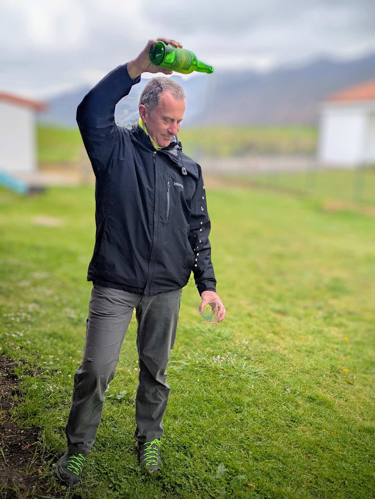 A man in a black jacket and gray pants stands in a grassy field, holding a green object above his head.