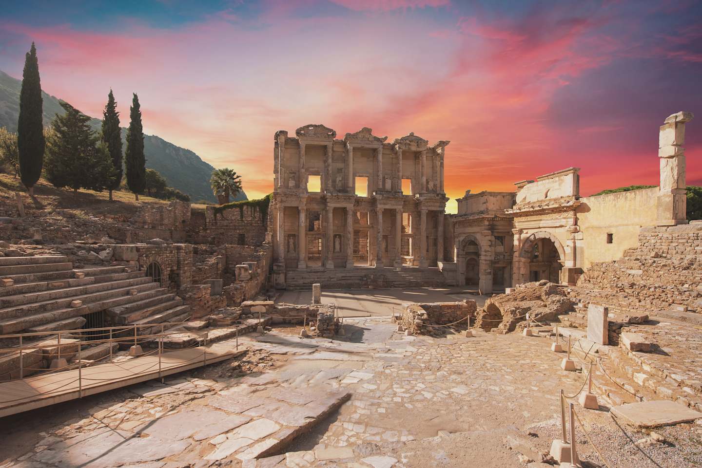 Majestic ancient ruins stand against a vibrant sunset sky, with steps leading up to the impressive structures in the foreground.