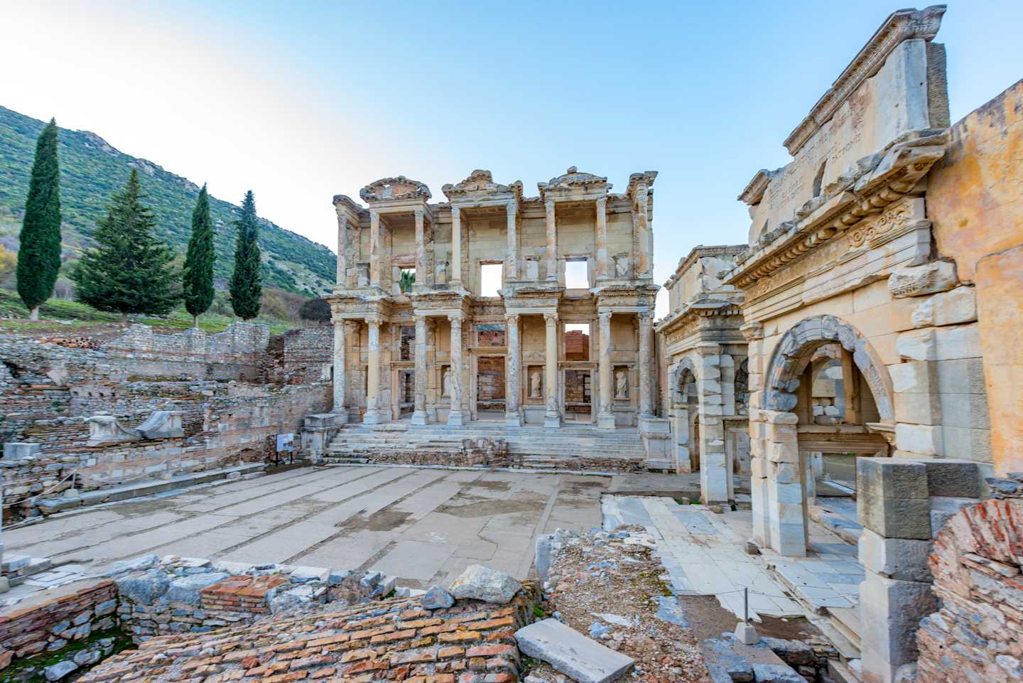 A grand, ancient library-like structure with ornate columns and arches stands amidst the ruins of an ancient city, surrounded by lush greenery and a mountainous backdrop.
