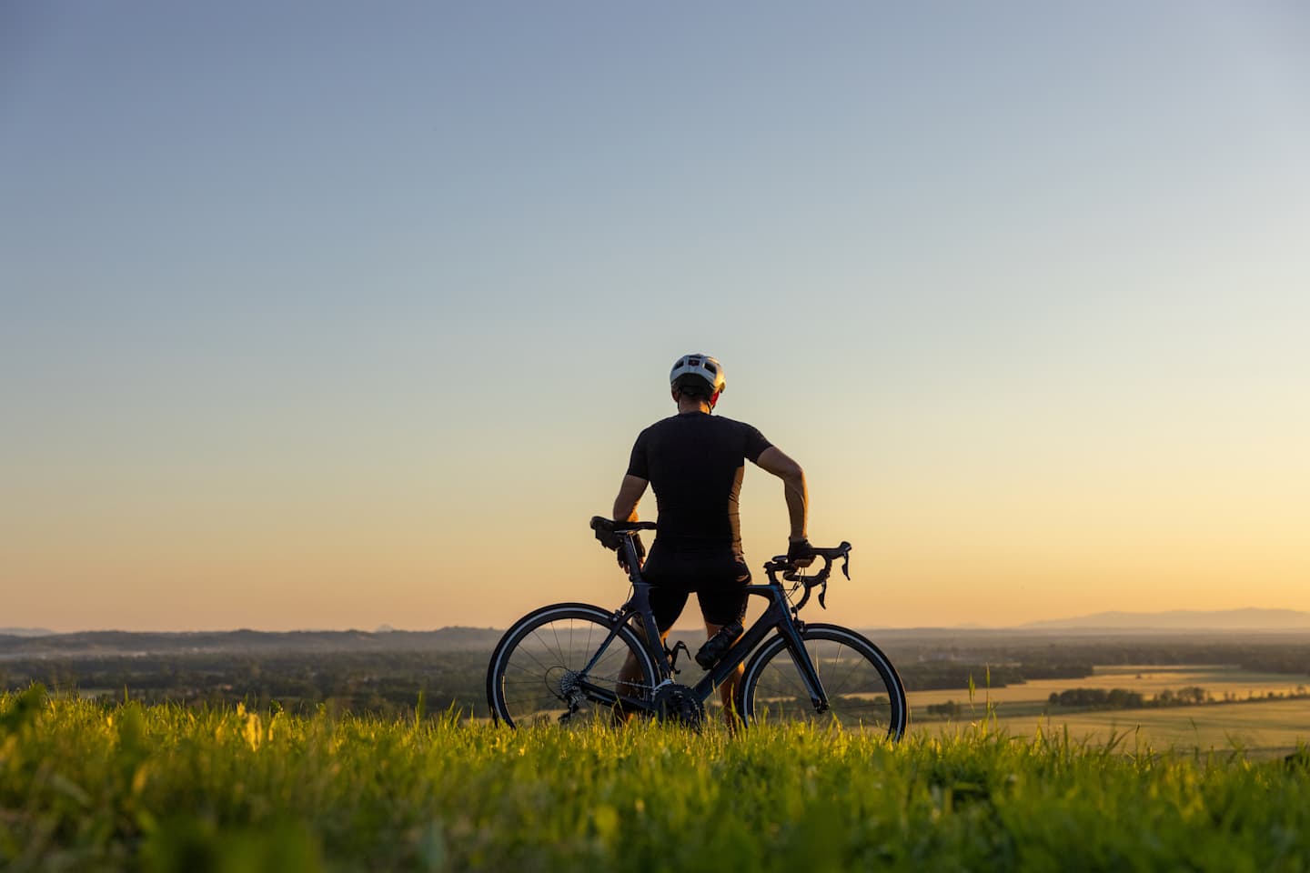 A cyclist stands on a grassy field, silhouetted against a vibrant sunset sky, with distant hills visible in the background.