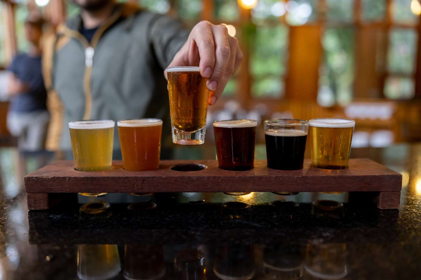 A wooden tasting flight with various craft beer samples, set against a blurred background of a rustic indoor setting.