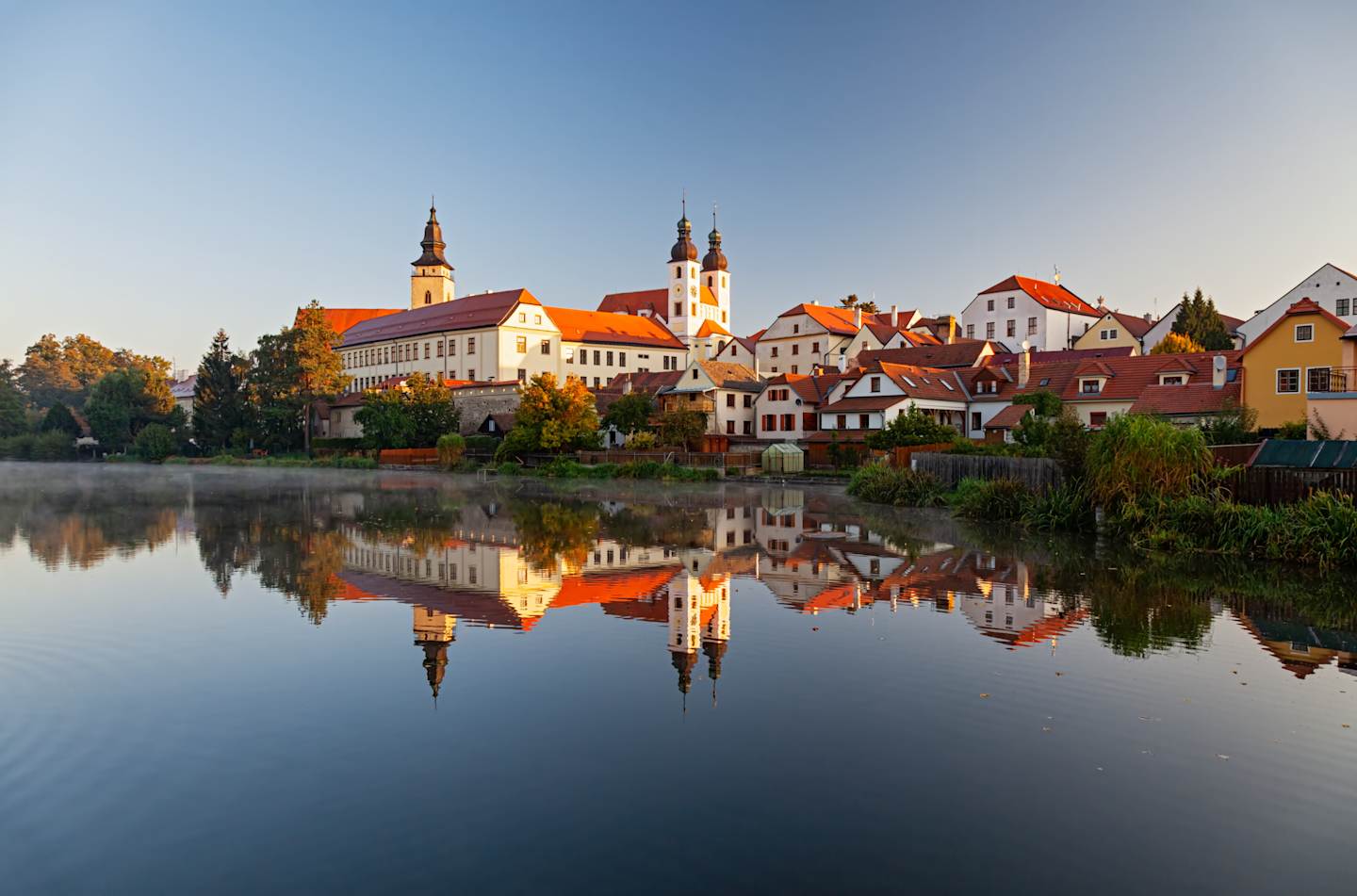 A picturesque town with colorful buildings and church spires reflected in the calm waters of a lake, set against a clear blue sky.