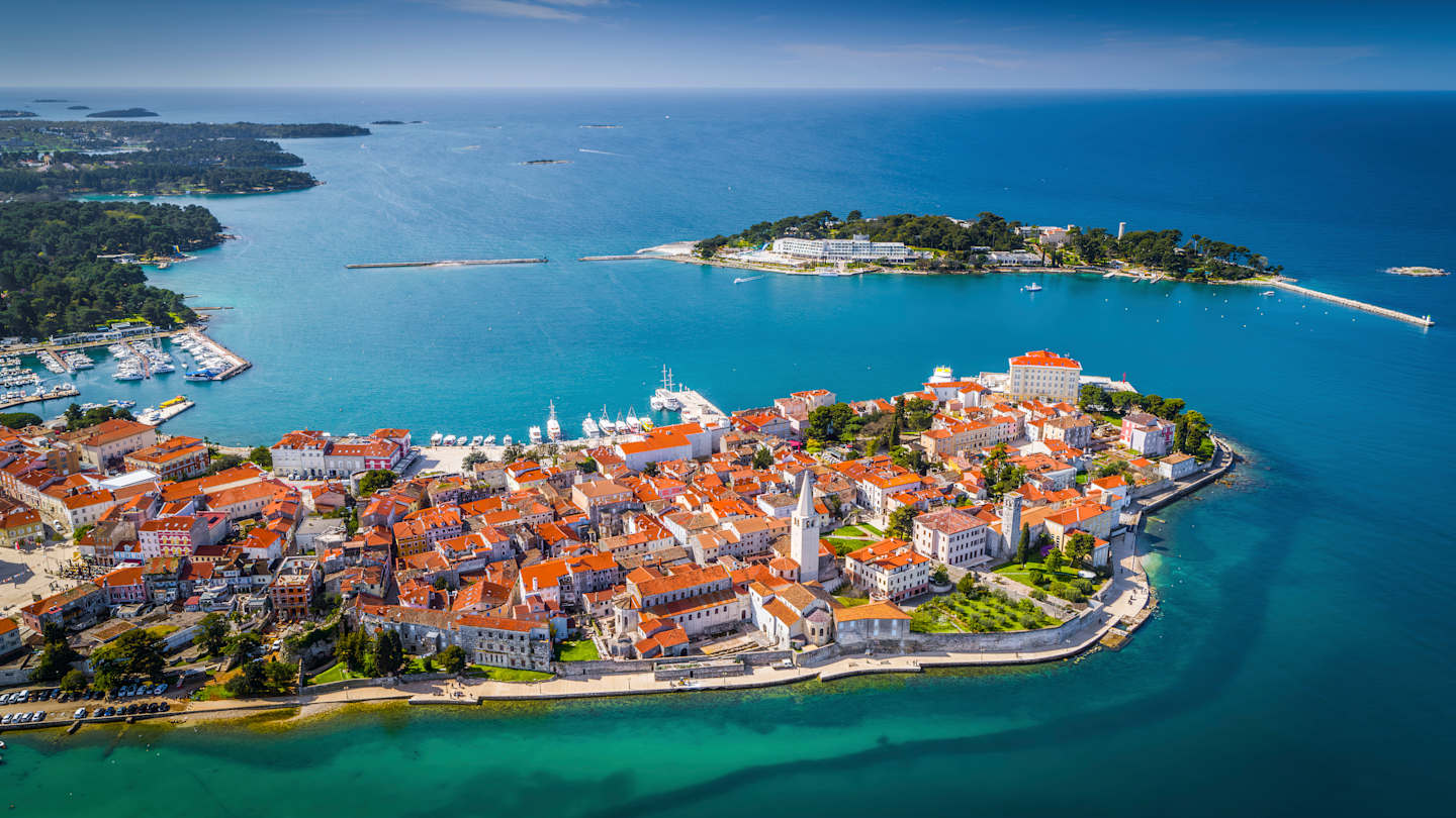 A picturesque coastal town nestled on a peninsula, surrounded by crystal-clear turquoise waters and dotted with vibrant orange-roofed buildings, with a lush green island in the background.
