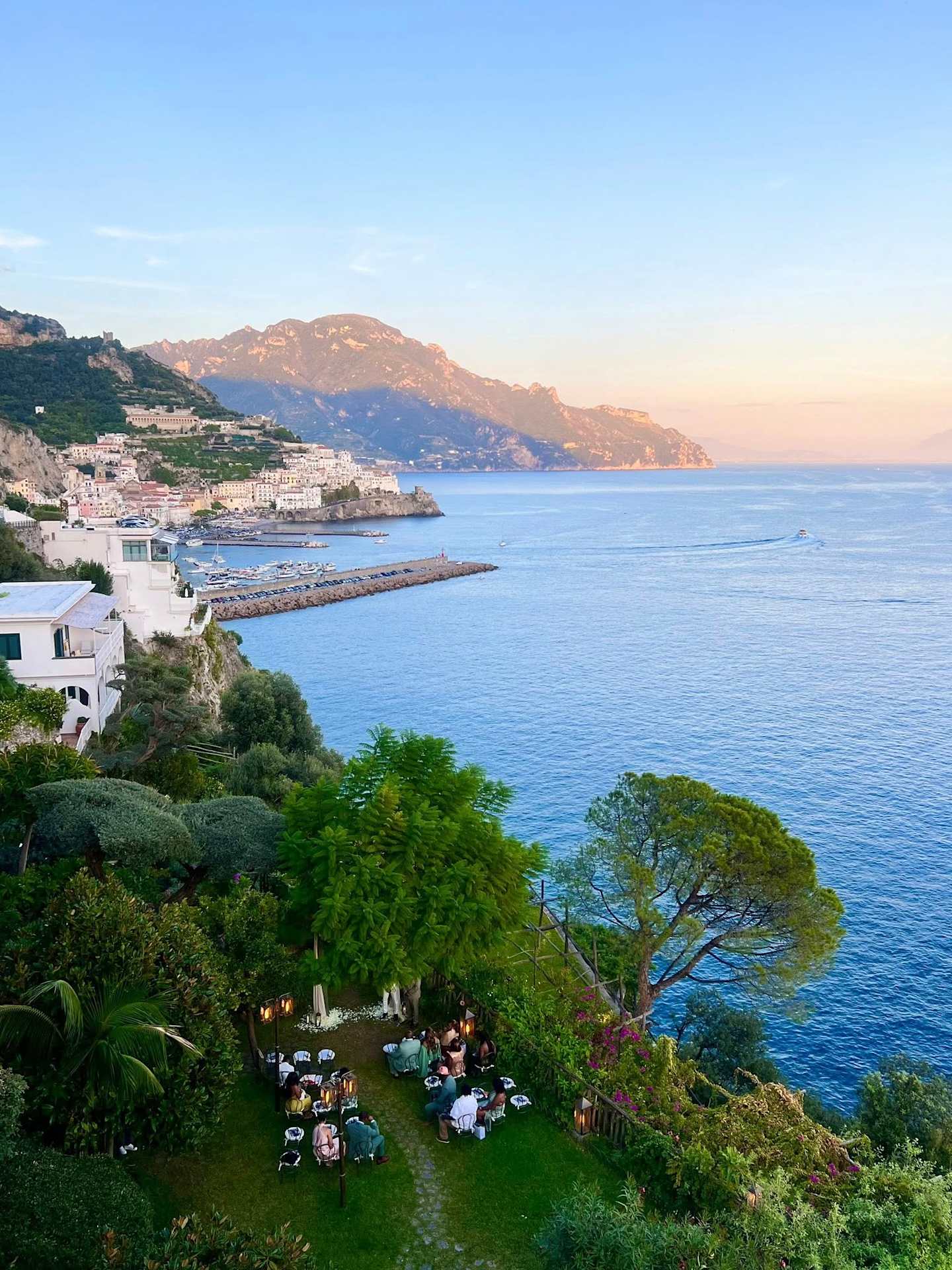 A picturesque coastal town nestled among lush greenery, with a scenic harbor and distant mountains visible under a clear blue sky.
