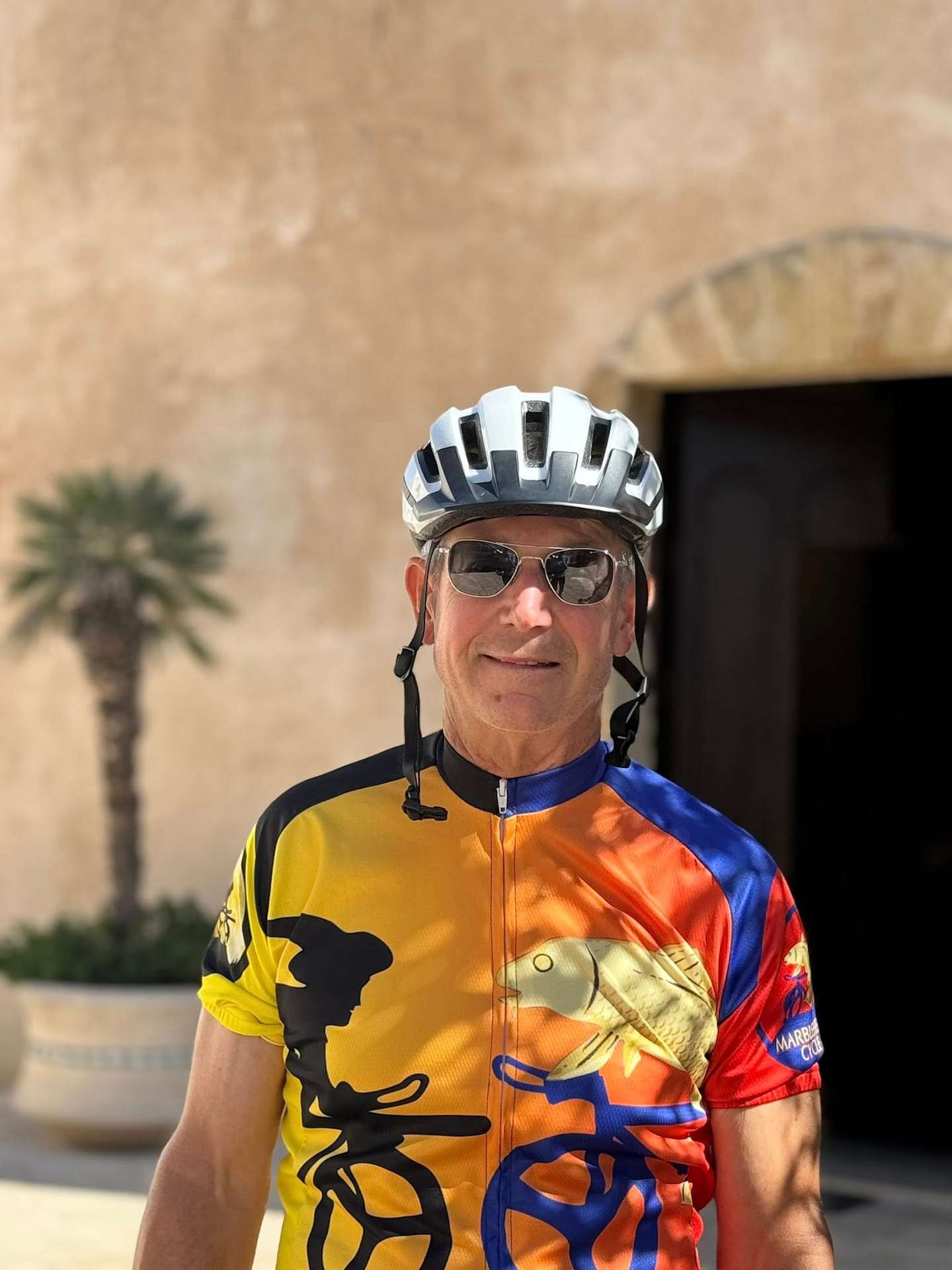 A person wearing a colorful cycling jersey and helmet, standing in front of a palm tree-lined background.