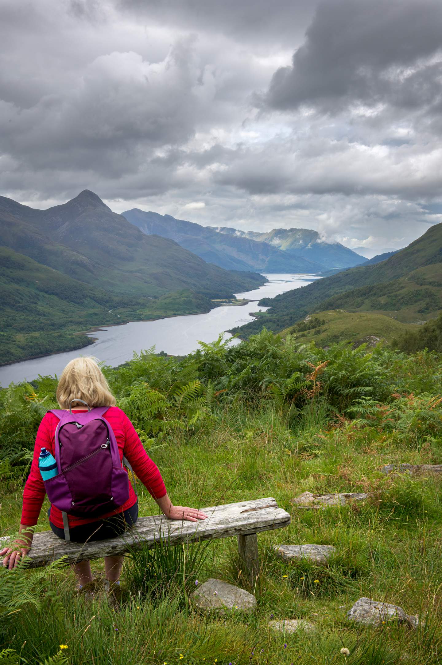 A person in a red jacket sits on a wooden bench overlooking a scenic landscape of mountains, lakes, and lush green vegetation under a cloudy sky.