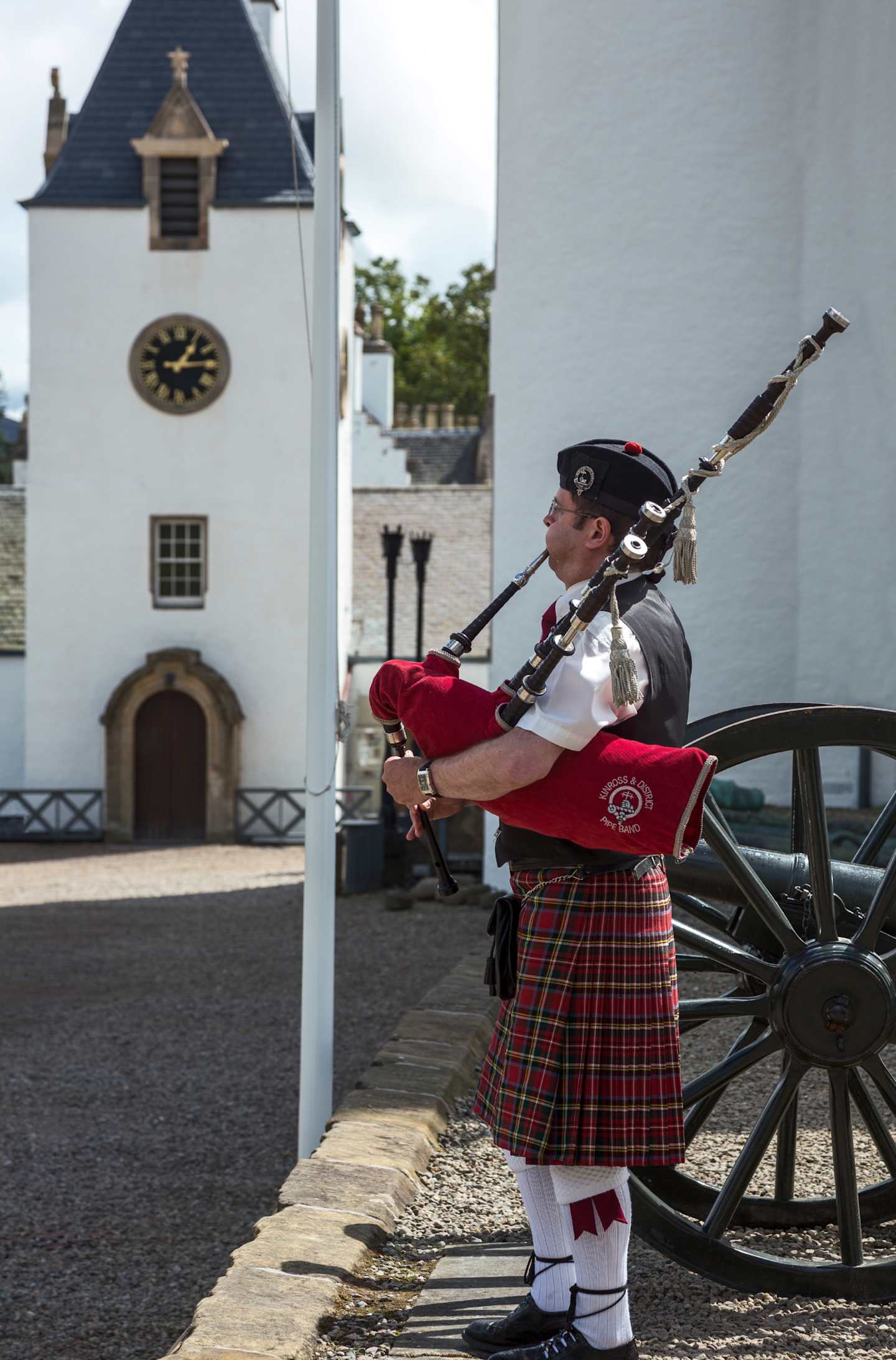 A person wearing traditional Scottish attire, including a kilt and playing the bagpipes, stands in front of a historic building with a clock tower in the background.