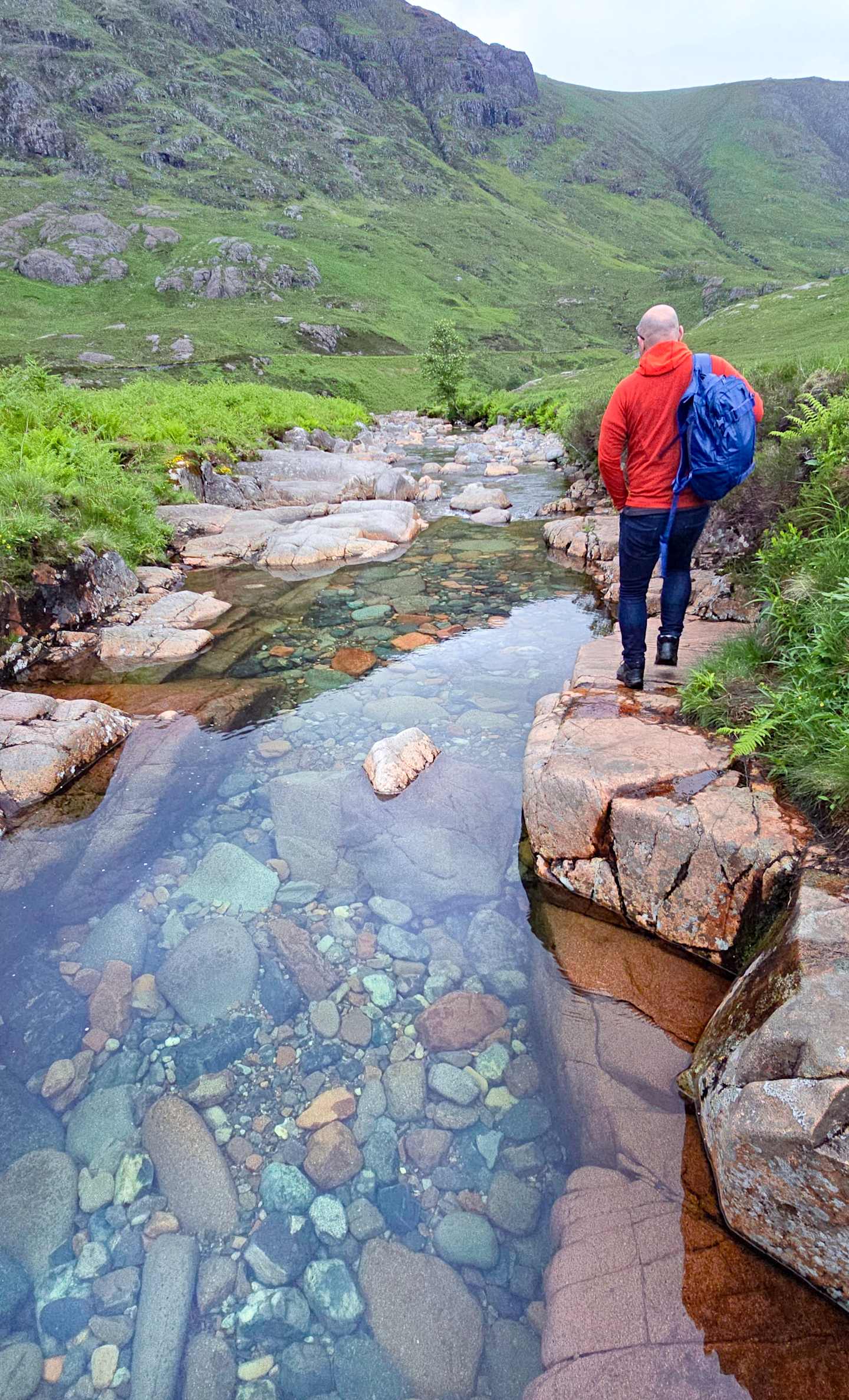 A person in a red jacket and blue backpack walks along a rocky stream in a lush, mountainous landscape.
