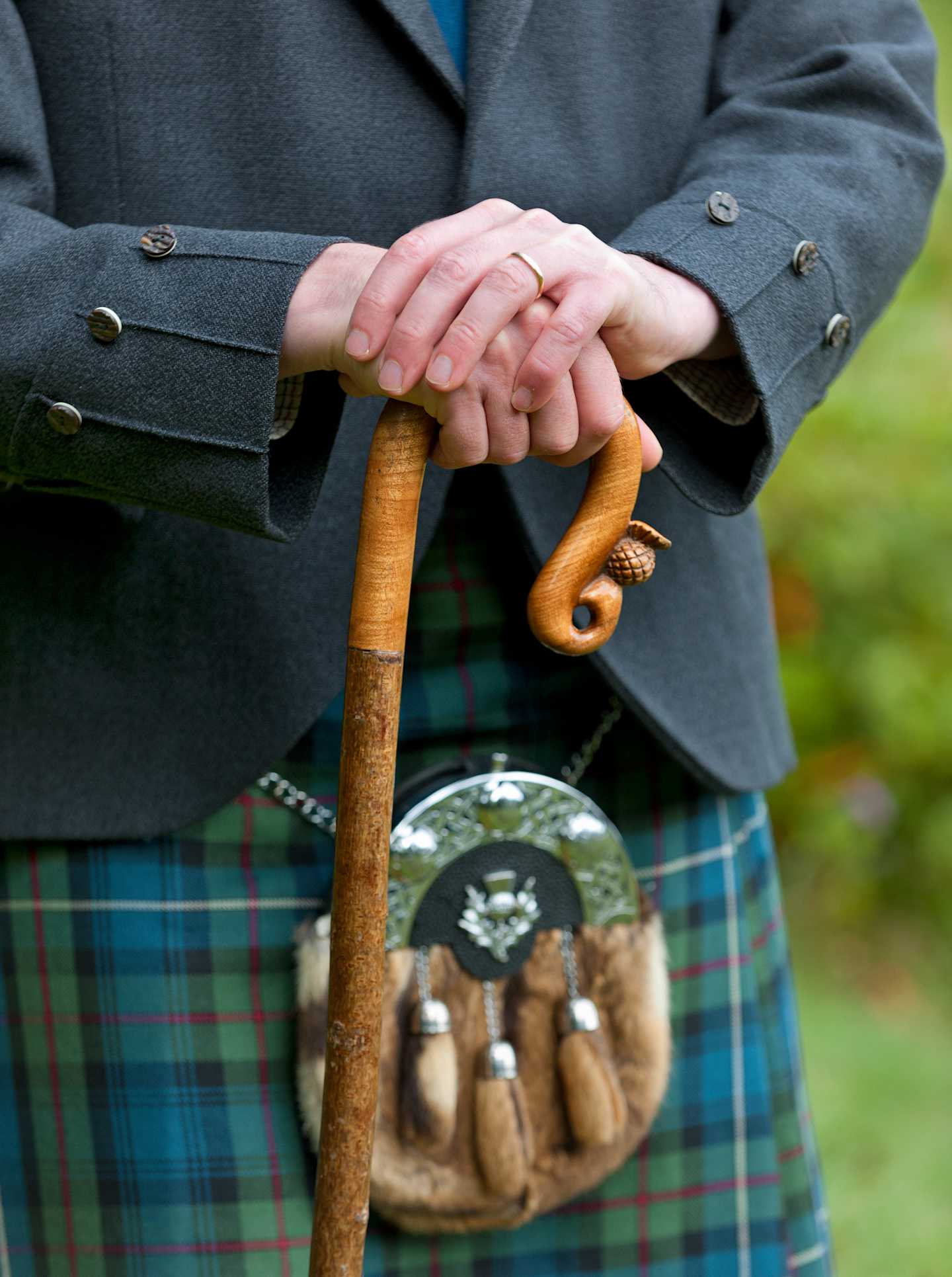 The image shows a person wearing a gray suit and holding a wooden walking stick, with a tartan kilt visible in the background.