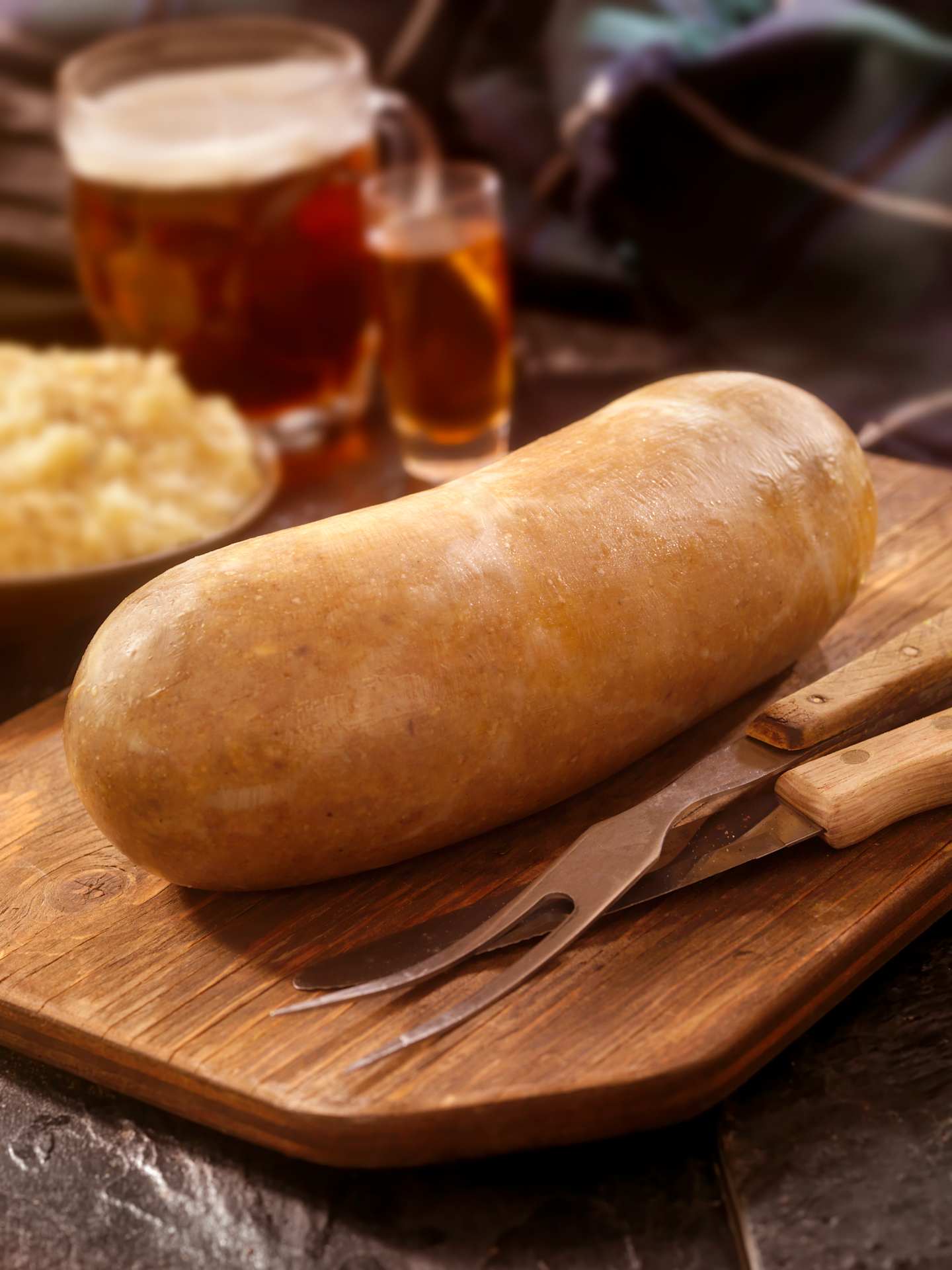 A wooden cutting board with a freshly baked bread roll, accompanied by glasses of amber-colored liquid, likely beer, and a plate of what appears to be sauerkraut or other fermented vegetables in the background.