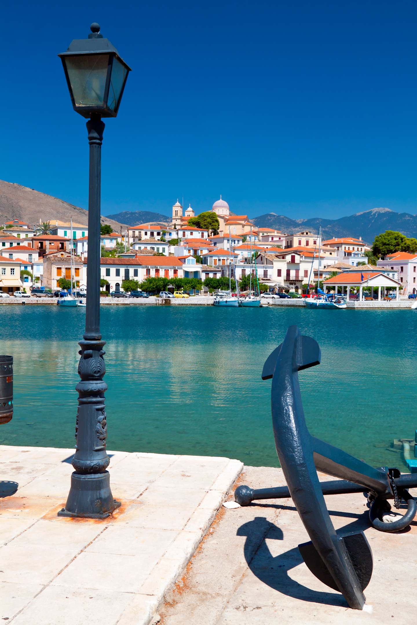 A picturesque harbor with a colorful town nestled in the mountains, featuring a decorative lamp post and an anchor in the foreground.