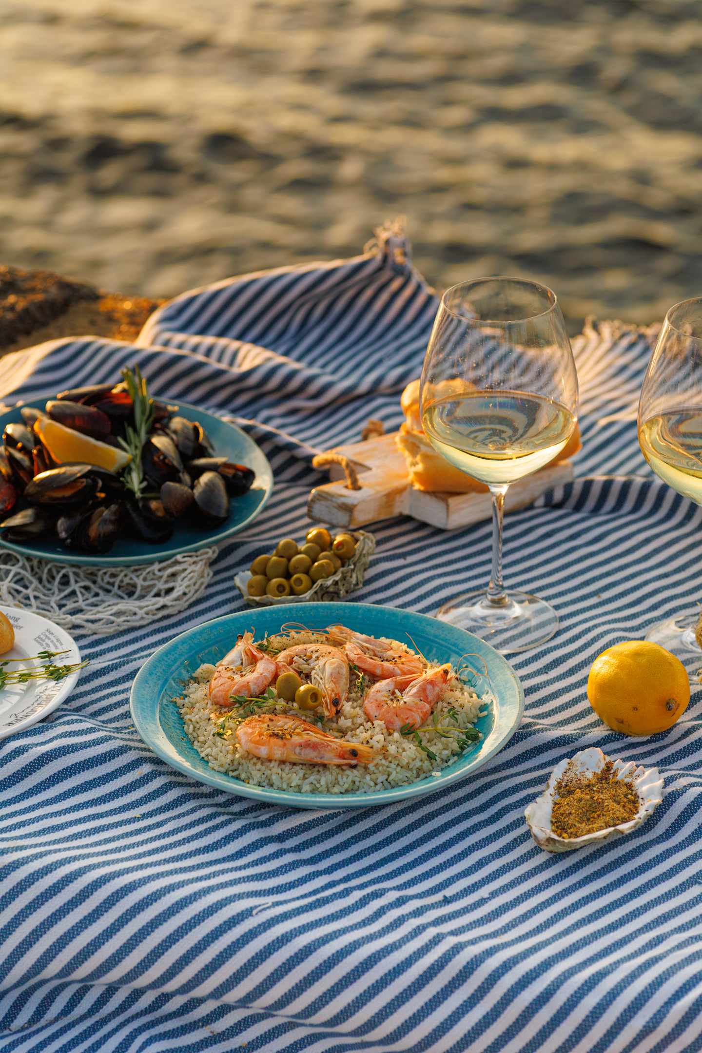 A picnic spread on a striped blanket by the water, featuring a plate of seafood and couscous, glasses of white wine, and a bowl of olives and other snacks.