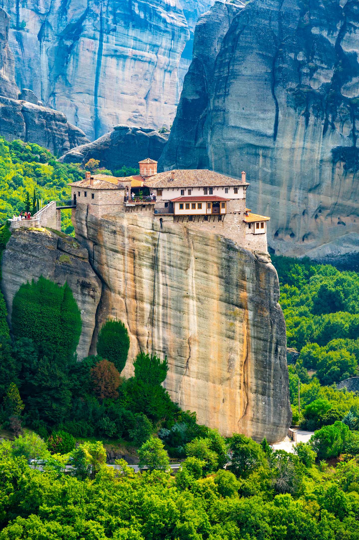 A stunning landscape featuring a towering rock formation with a monastery perched atop, surrounded by lush, verdant vegetation in the foreground.