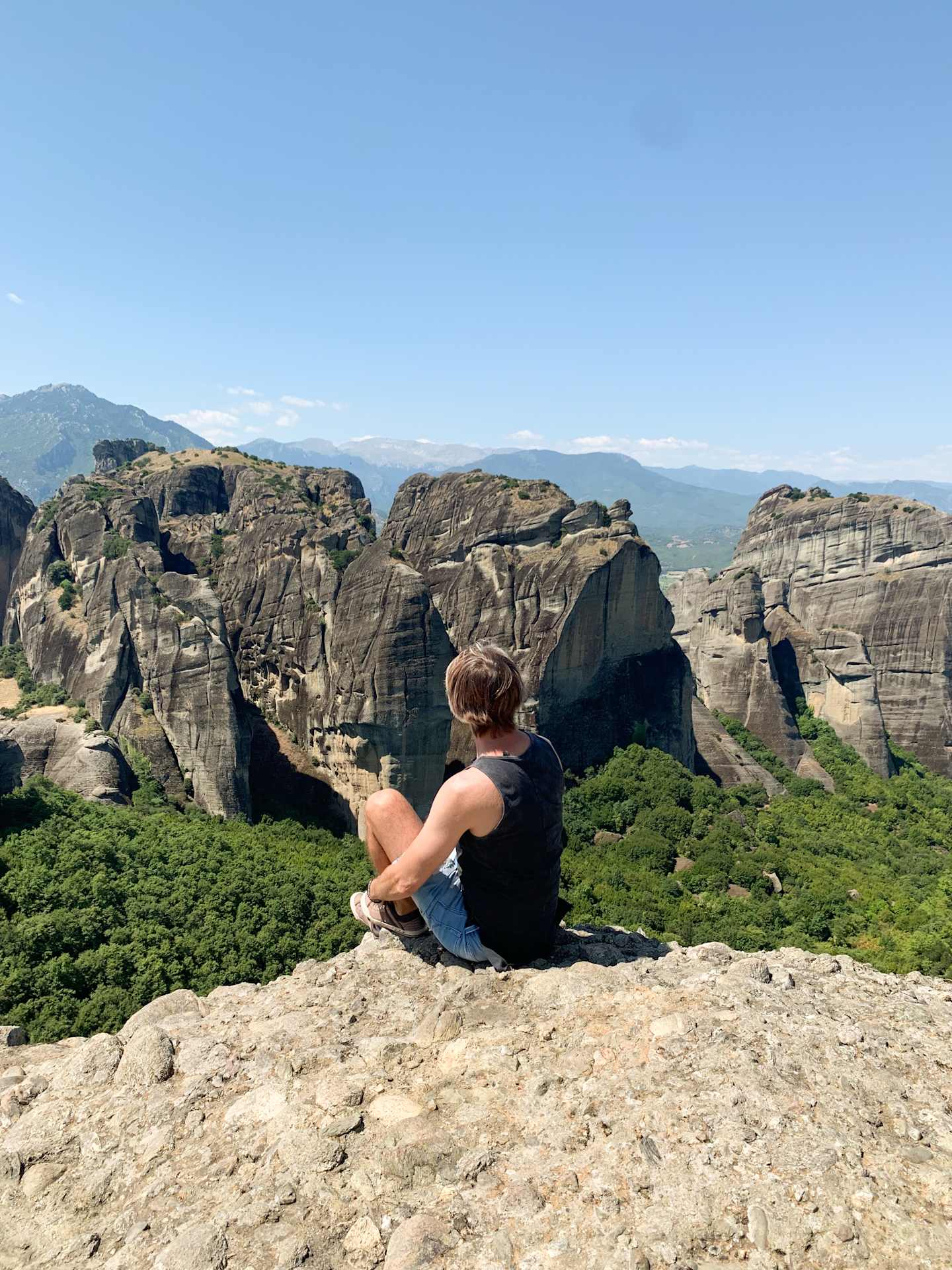 A person sitting on a rocky outcrop overlooking a scenic landscape of rugged mountains and lush vegetation in the background.