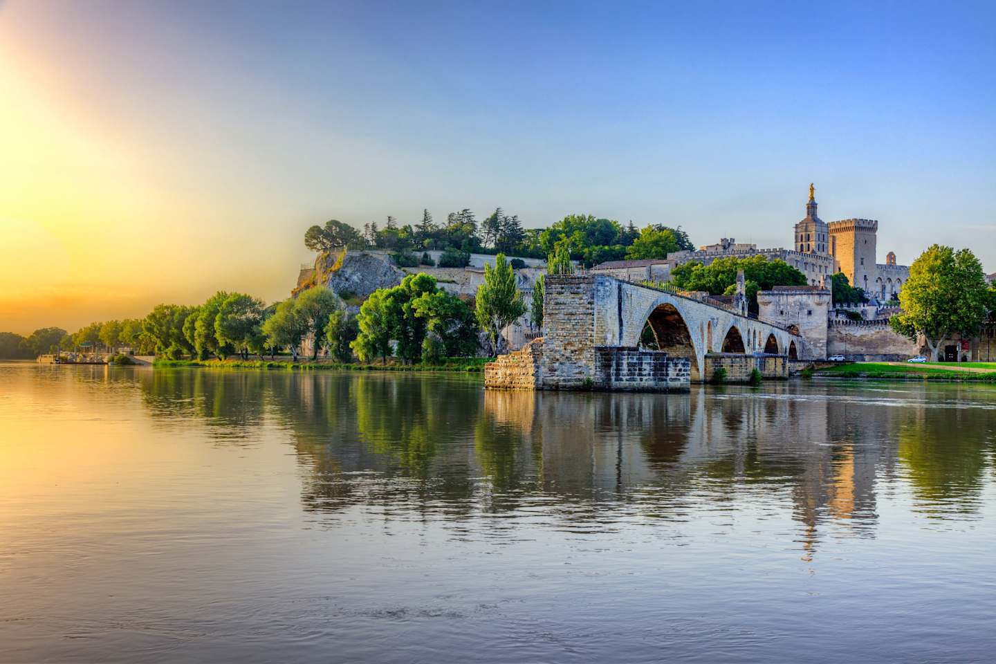 A picturesque medieval bridge spans a tranquil river, with a historic town nestled on the hillside in the background, bathed in the warm glow of the setting sun.