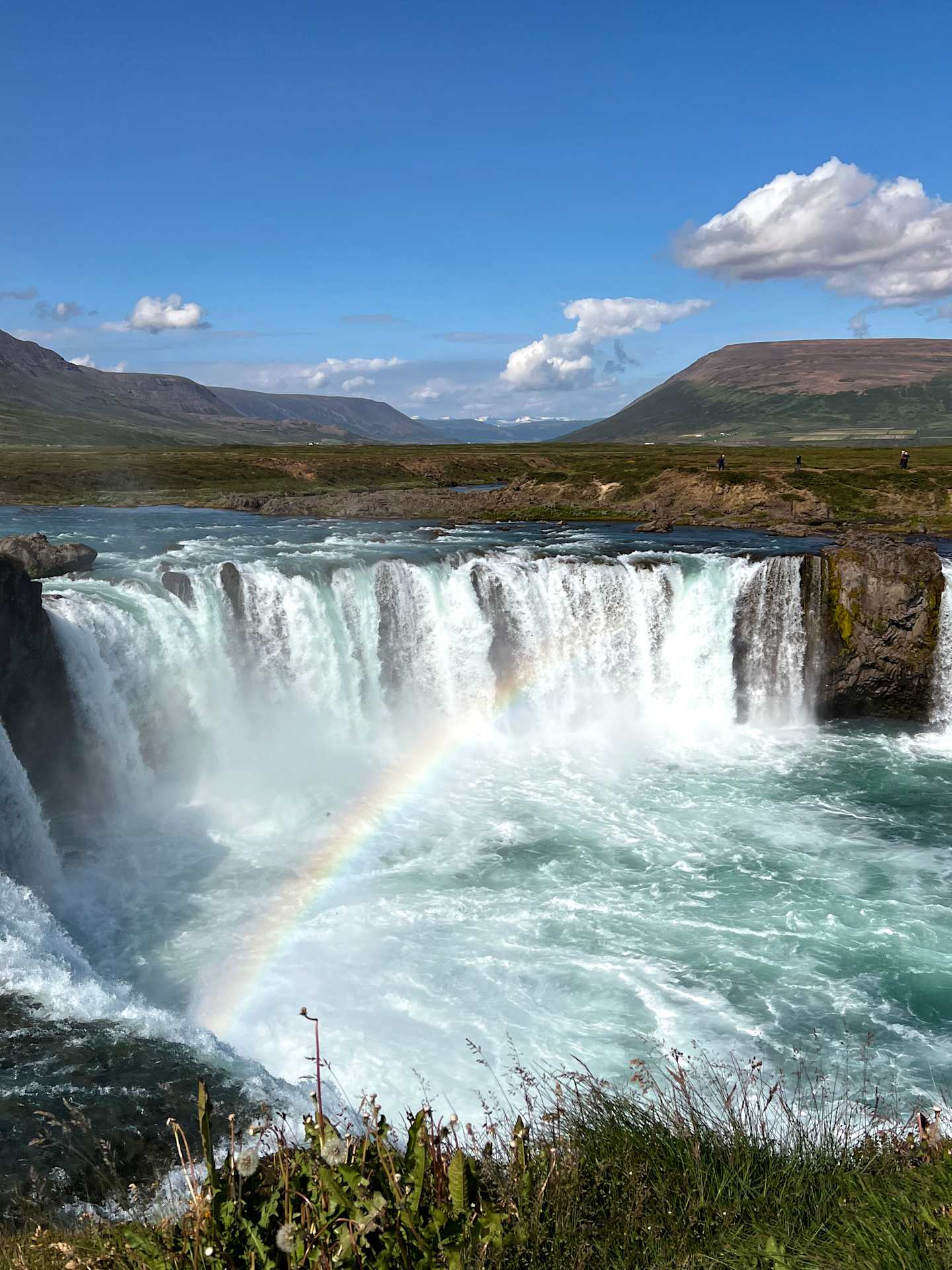 A majestic waterfall cascades over a rocky cliff, creating a stunning display of power and natural beauty, with a vibrant rainbow arching across the misty spray in the foreground, set against a backdrop of rugged mountains and a clear blue sky.