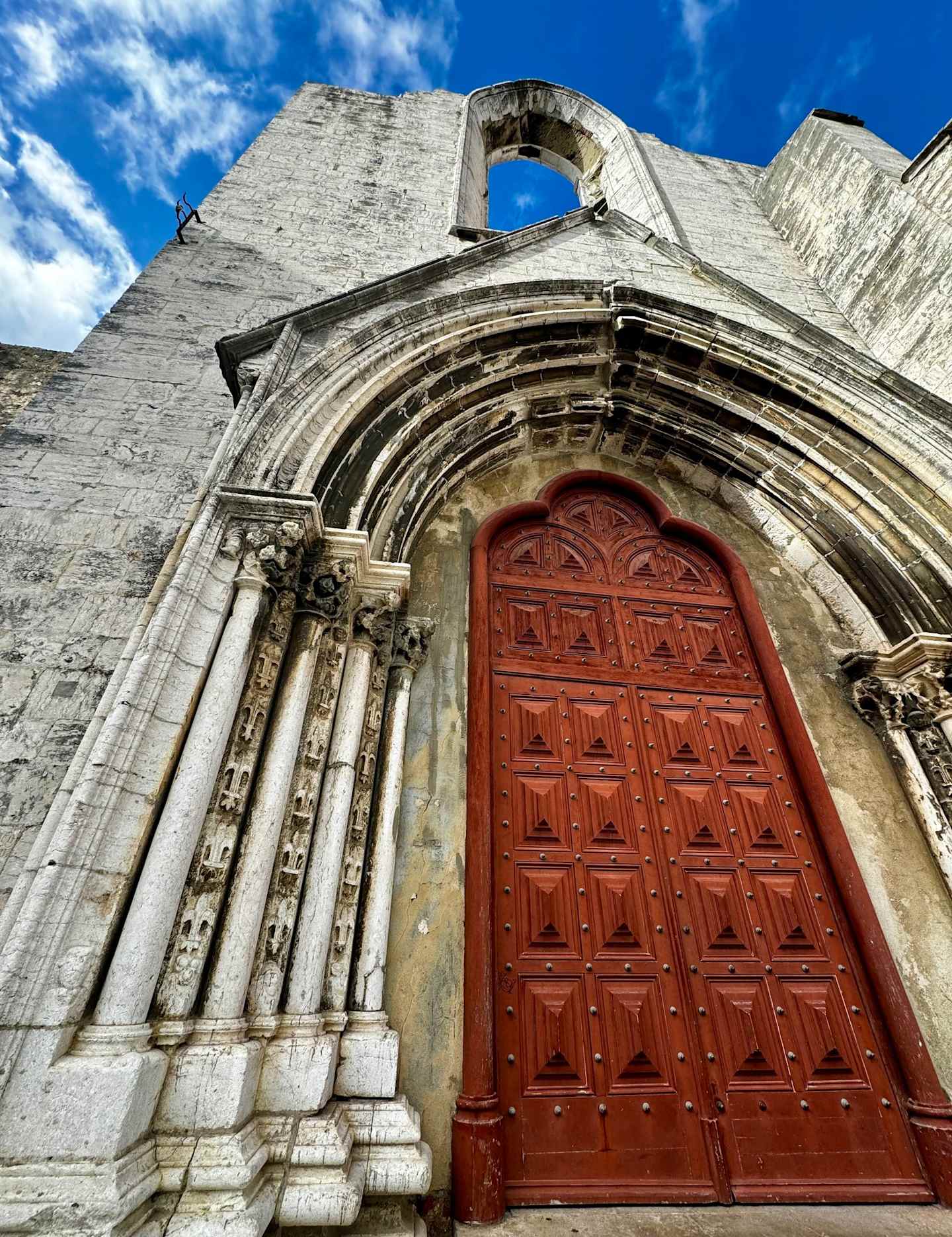 A grand, ornate wooden door set within an arched stone entryway, framed by a blue sky with fluffy white clouds.