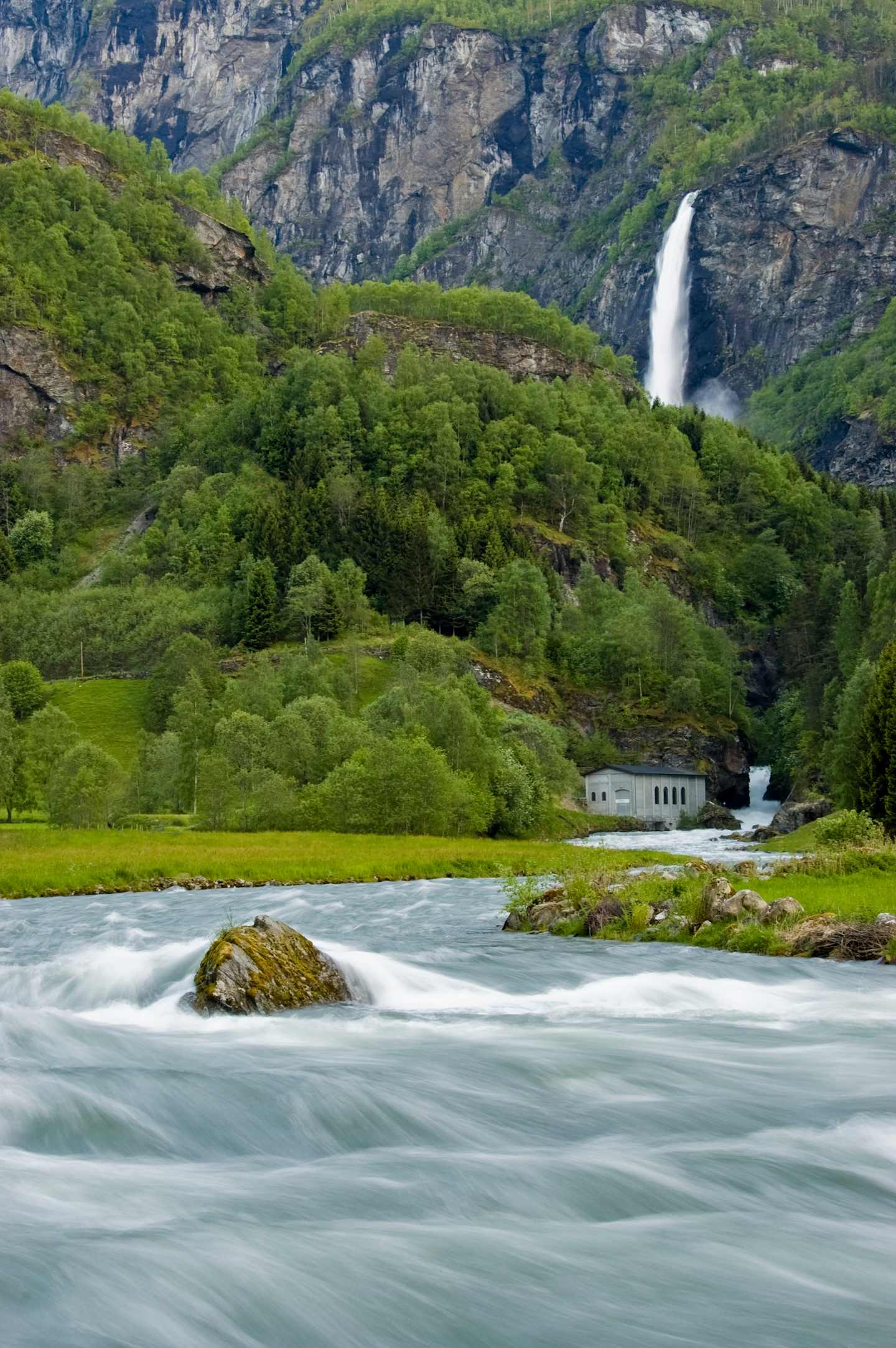 A lush, green landscape with a cascading waterfall, a flowing river, and a small building nestled among the vegetation, all set against a backdrop of rugged, mountainous terrain.