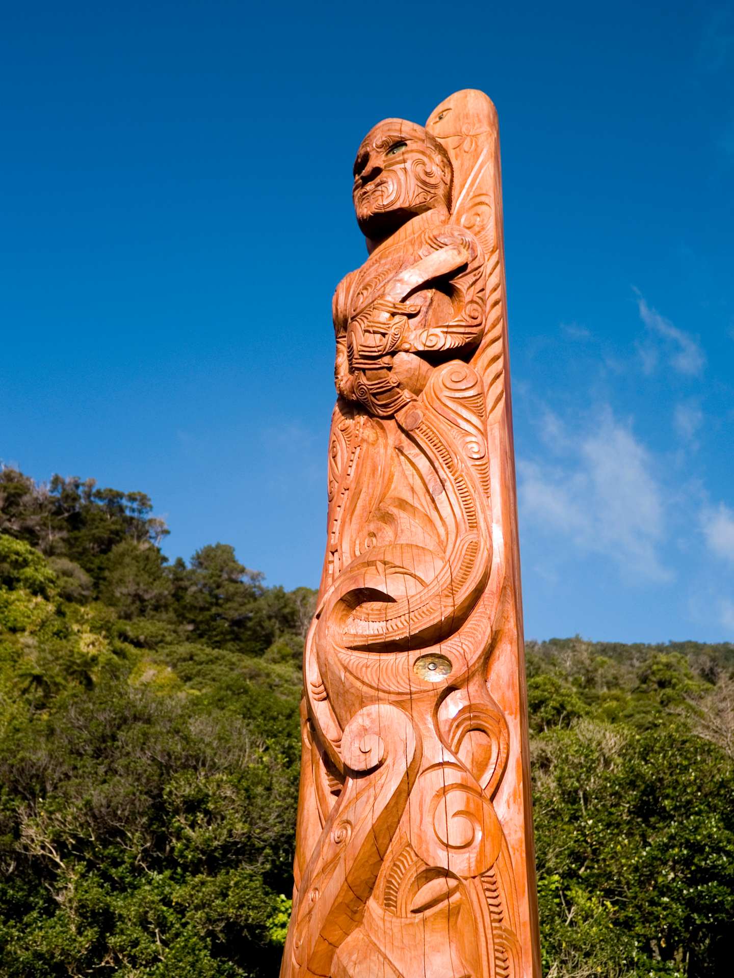 A large, intricately carved wooden sculpture of a figure stands prominently against a backdrop of lush greenery and a clear blue sky.