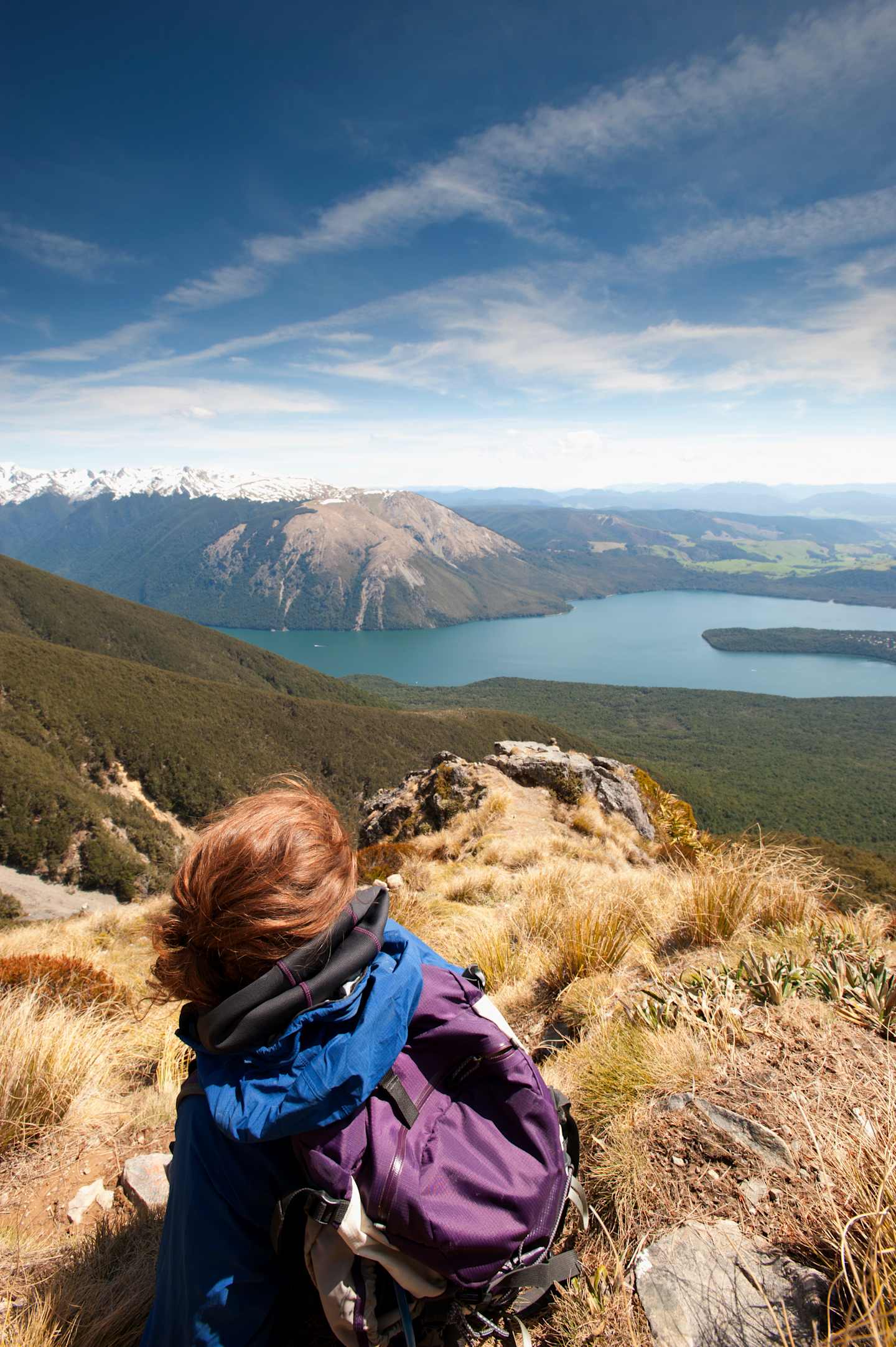 A person wearing a backpack sits on a rocky outcrop overlooking a vast, scenic landscape with a large lake and snow-capped mountains in the distance.