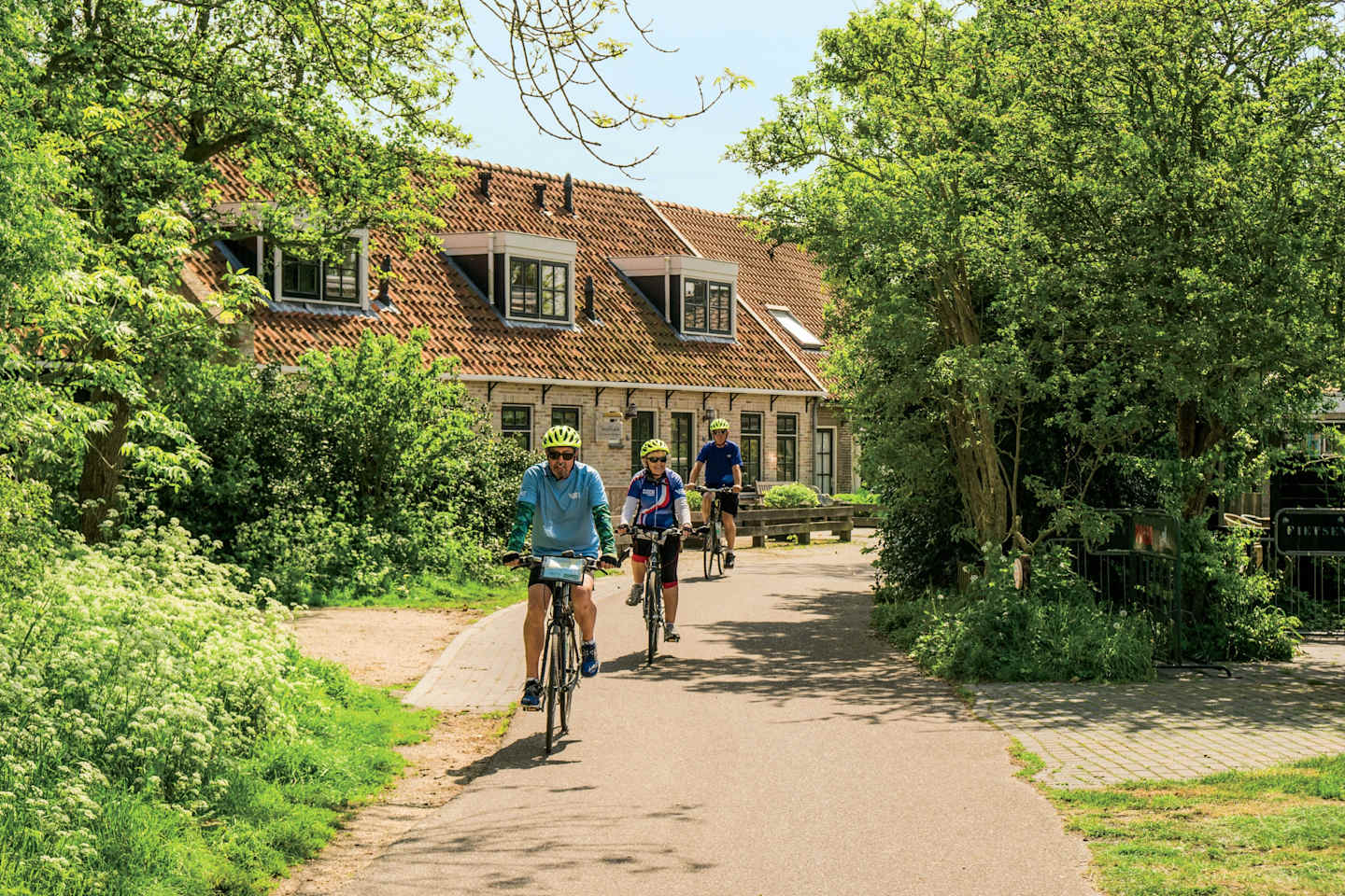A scenic path winds through a lush, green landscape, with a charming building and cyclists enjoying the outdoors in the background.