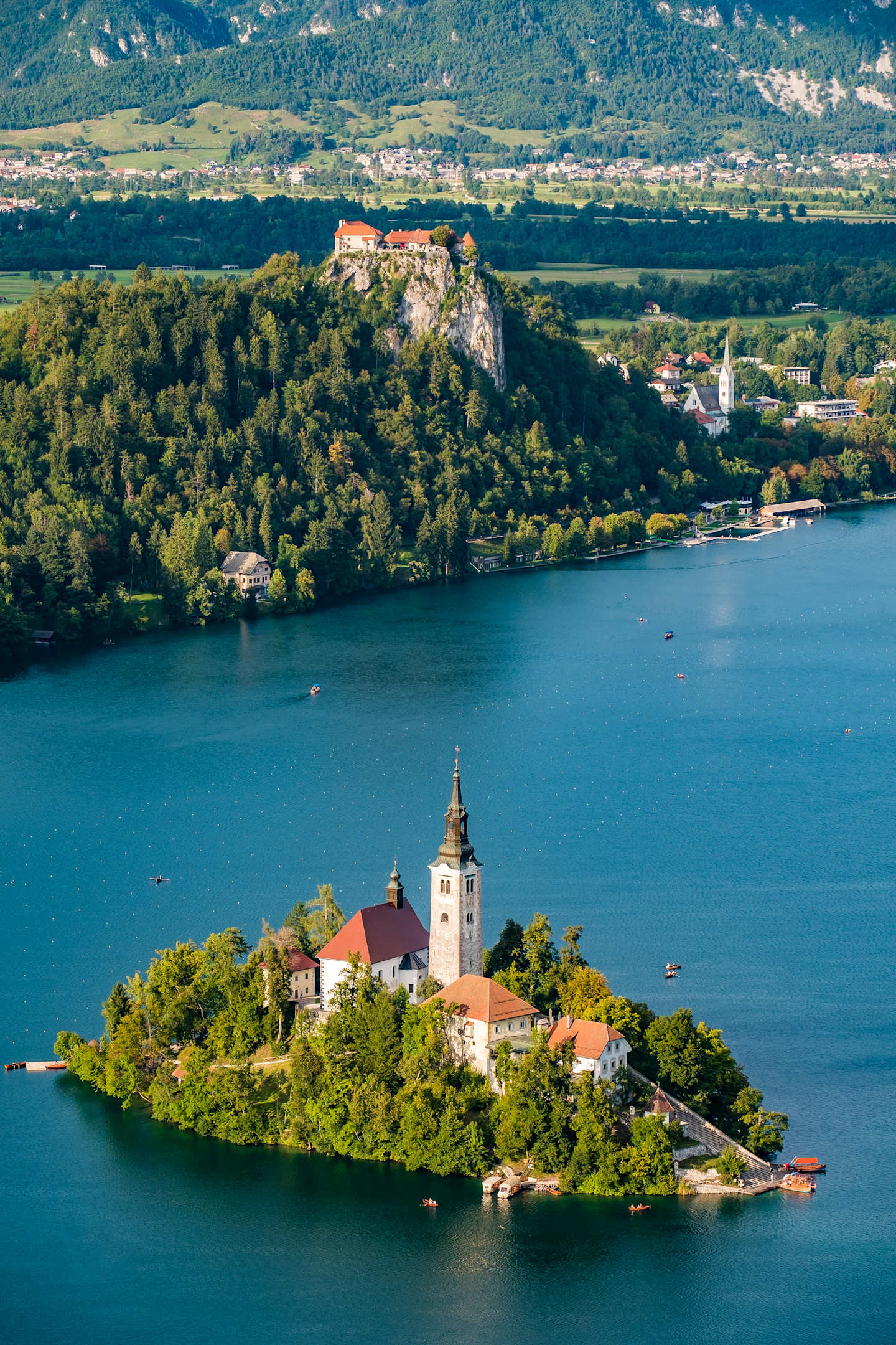 A picturesque island with a church and buildings nestled among lush greenery, set against a backdrop of mountains and a serene blue lake.
