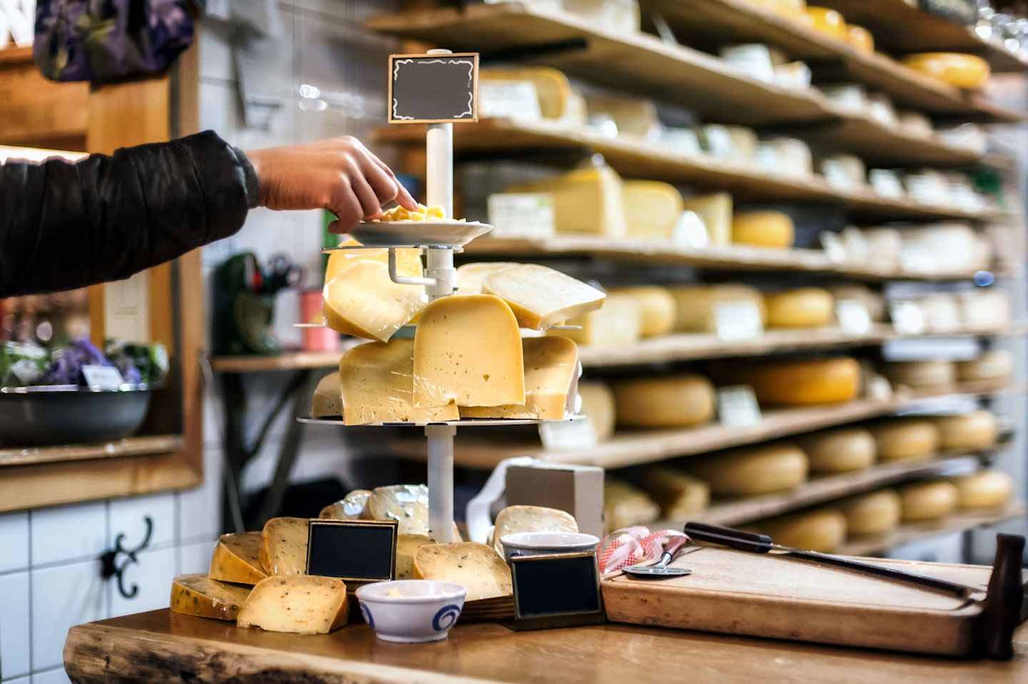 A hand reaching to select a piece of cheese from a display of various cheeses on shelves in a store or market.