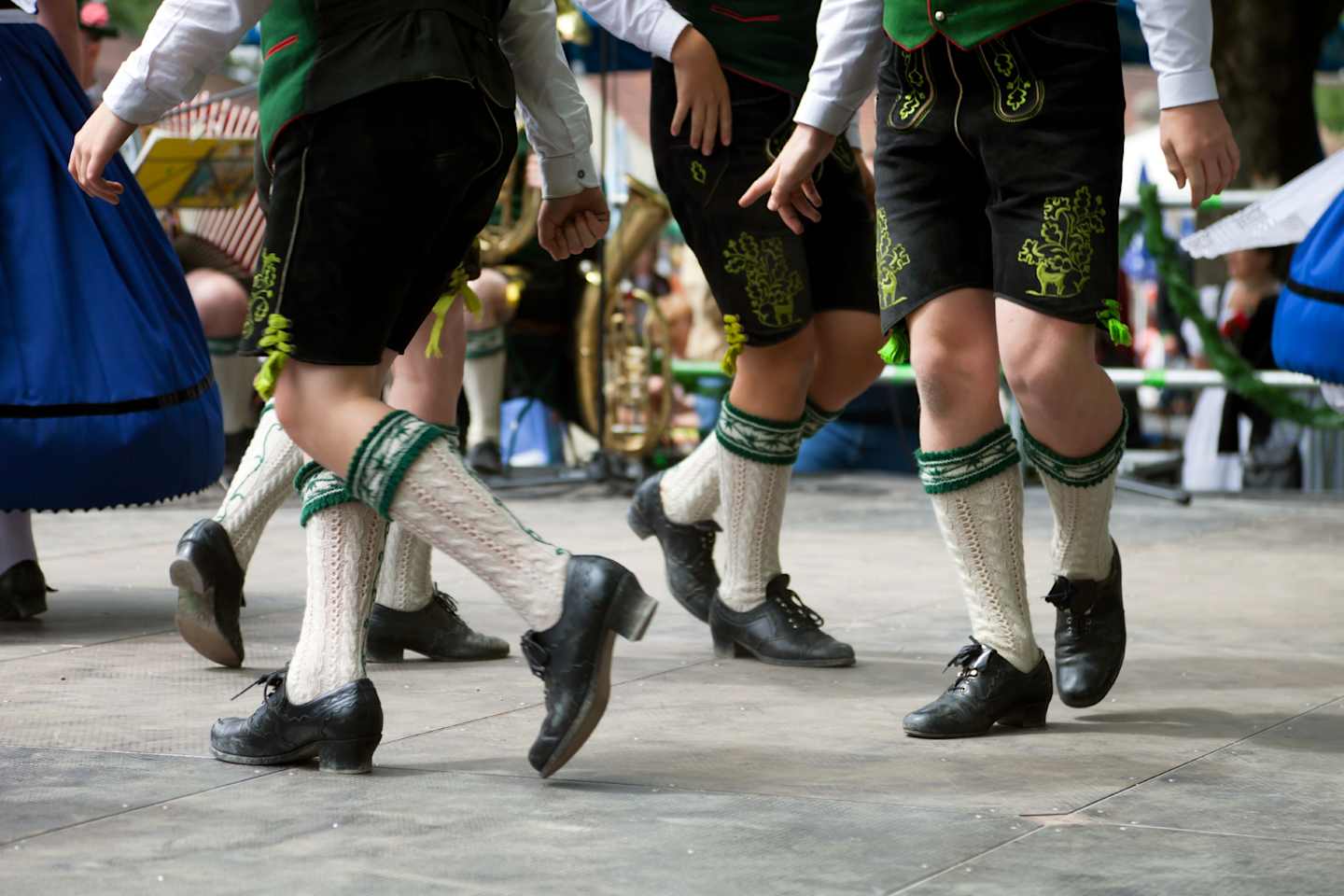 The image shows the lower bodies and feet of people wearing traditional Irish dance costumes, including black shoes with hard soles, white stockings, and green and black patterned skirts. The background appears to be a stage or performance area.